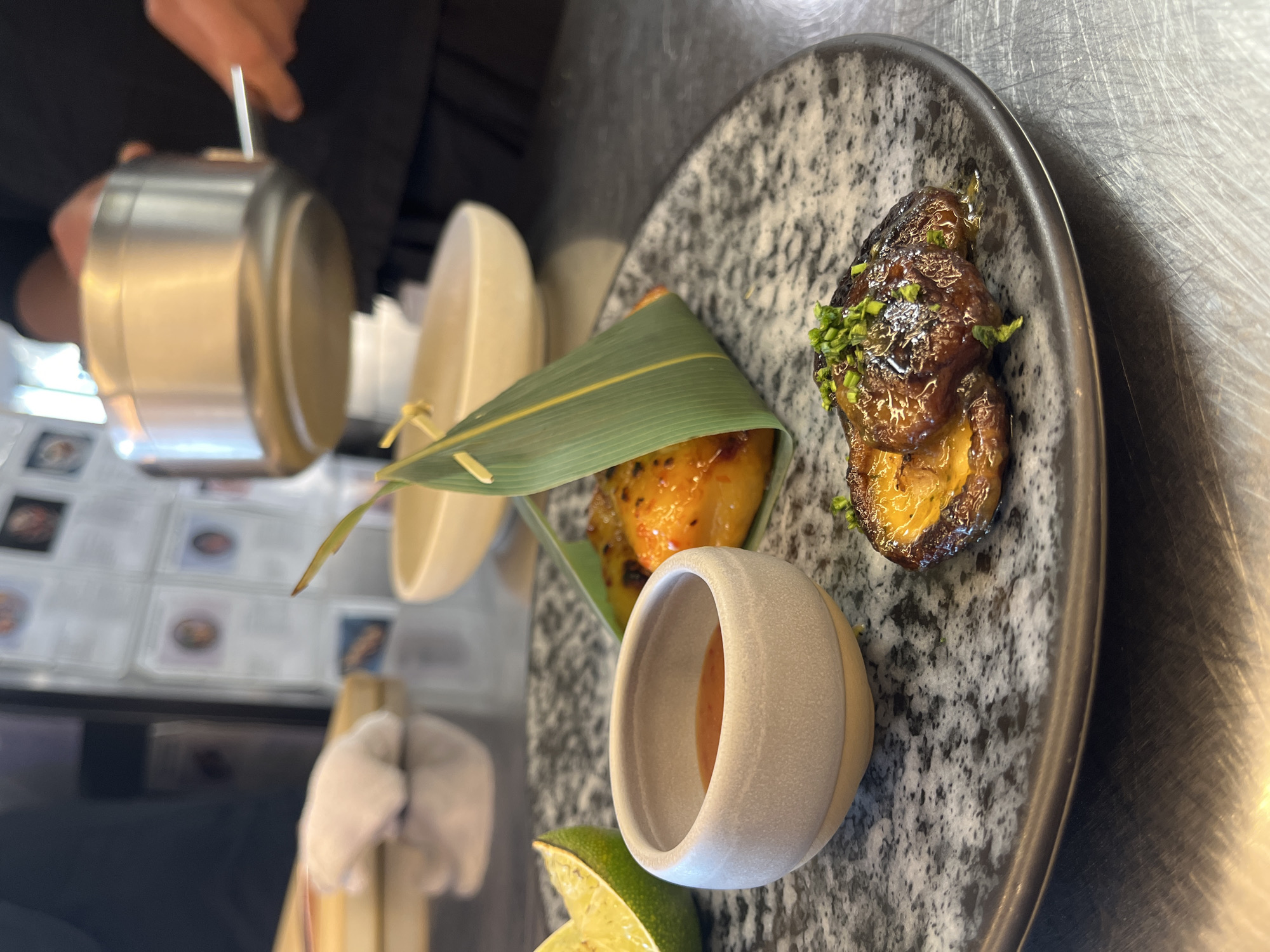 The image shows a plate of food being prepared in what appears to be a professional kitchen setting. The plate is a speckled grey and black, featuring several distinct elements: a cone-shaped object wrapped in a green leaf, an individual small bowl with a sauce or dip inside, and a portion of glazed and garnished meat or vegetable. A halved lime is partially visible at the edge of the plate. A person is holding a silver pot with what looks like liquid inside, seemingly preparing to add it to the dish.
Behind the plate, there is a background with more professional kitchen equipment and a menu with portraits. The scene is indoors. The exact location within Sofia, Bulgaria is not determinable from the image alone.