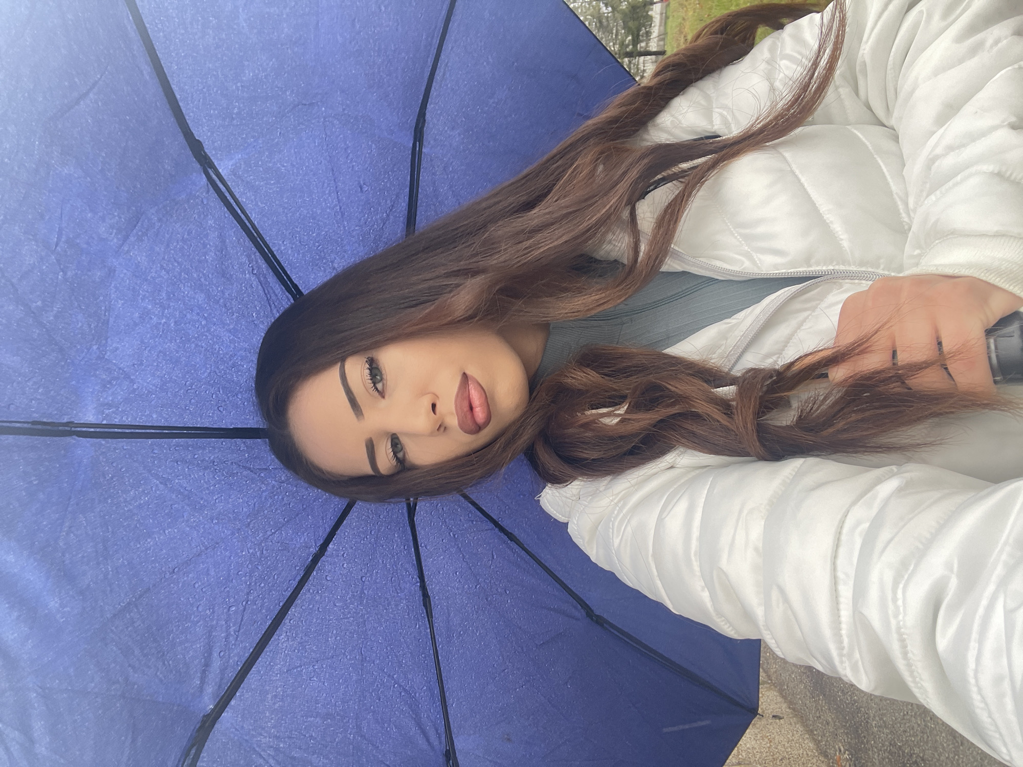 A young woman poses for a photo under a blue umbrella speckled with raindrops. She is wearing a white, quilted jacket over a light-colored top. Her long, brown hair is styled in loose waves. The background is blurry, but appears to be an outdoor location with some green foliage. This image does not contain sufficient information to identify the precise location in Veliko Tarnovo, Bulgaria.