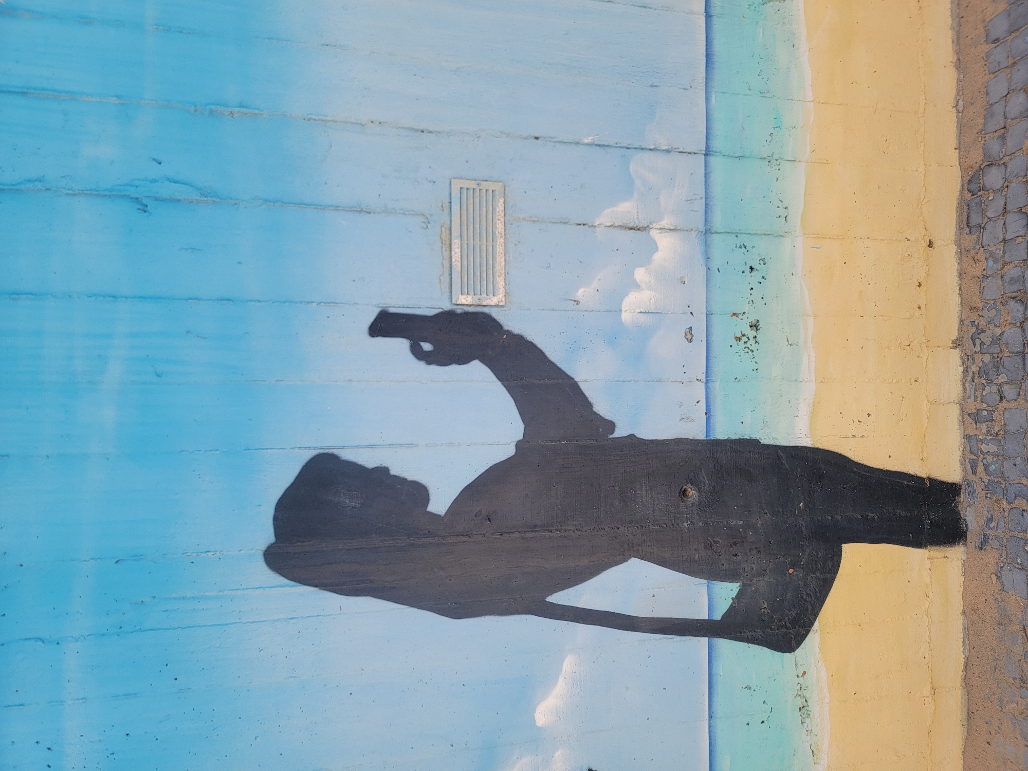 A mural depicts a silhouette of a person taking a selfie on a beach. The mural is painted on a concrete wall, with the upper portion featuring a light blue sky with white clouds, transitioning to a turquoise ocean and a yellow sand beach. The silhouette is that of a person with their hair pulled back in a ponytail, holding a smartphone in their right hand. A dark bag hangs from their left shoulder. In the background, on the wall, is a rectangular ventilation grille. The foreground shows a cobblestone pathway. The mural is located on the promenade of Quarteira, Portugal.