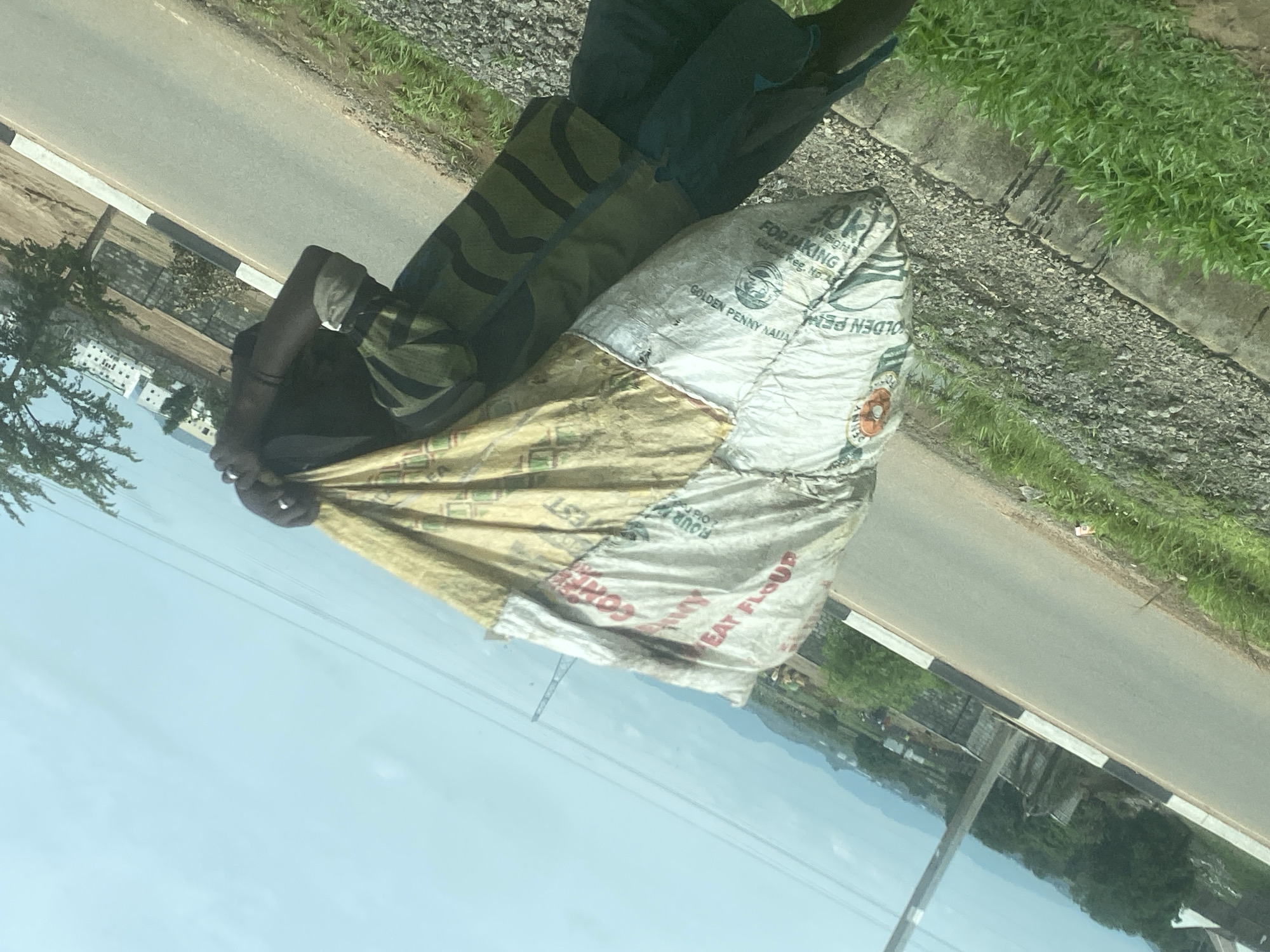 A vertical, eye-level shot captures a person carrying large sacks by the side of a road, in what appears to be Gosa, Nigeria. The individual, seen from the chest down, wears a dark shirt and camouflage-patterned trousers. They are holding two large sacks: one white and one light brown/yellow.
The white sack clearly displays text such as "FOR BAKING", "GOLDEN PENNY NAIJA", "RAFDAC Reg. No.A", and "Weight", along with partially visible words like "EAT FLOUR" and "FOUR" (some text is upside down). The light brown/yellow sack has fragmented text including "EST", "OLD", and "CORN". These texts suggest the sacks contain food products, likely flour and corn, from a Nigerian brand.
The scene unfolds outdoors on a bright, clear day, indicated by the light blue sky and adequate natural illumination. In the background, a paved road with a black and white striped curb runs alongside a gravel shoulder and green grass. Beyond the road, there are trees, some distant buildings, and what appears to be hills or a densely vegetated area. Power lines are visible against the sky. The perspective suggests the photo was taken from a vehicle.
