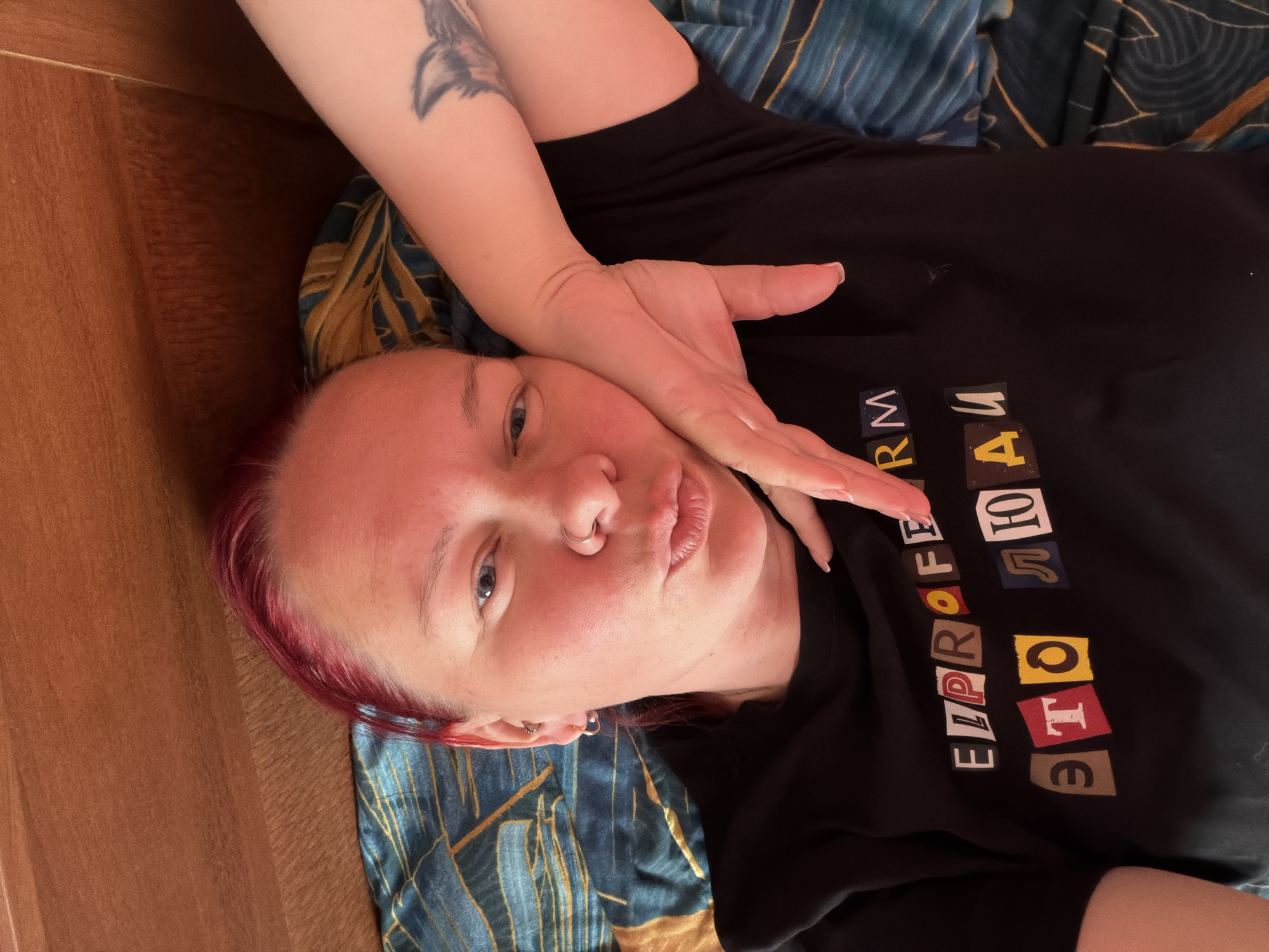 A fair-skinned person with short, dyed-red hair is lying on a surface that appears to be part wood (possibly a wall) and part blue and gold fabric (possibly bedding), with the person facing the camera and partially obscured by their own hand. The person's right hand is resting on their face, covering part of their cheek. The person has blue eyes, a nose ring, and an earring visible in their left ear. On the person's left arm, which is raised behind their head, is a tattoo of a bird. The person is wearing a black t-shirt with the word "ELPROFOTE" in Cyrillic script (ЭЛПРОФОТЕ), written in block-letter style with letters constructed as if from magazine cutouts.
The location is not discernible from the image.