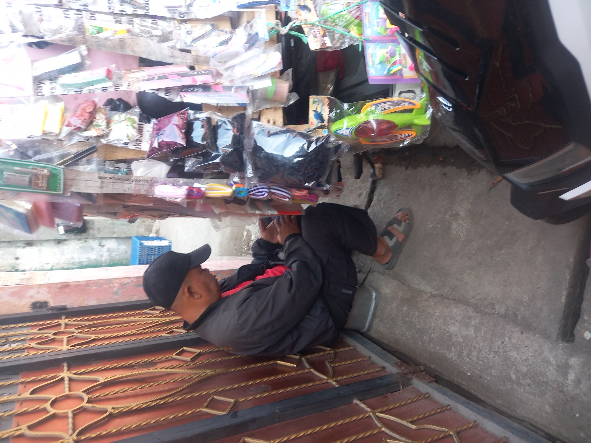 In Linggar, Indonesia, an outdoor scene shows a man wearing a black baseball cap and a black jacket over a red shirt, sitting on a grey stool. He is looking down at his hands, which appear to be holding a dark-colored smartphone. The man is positioned beside a closed gate with intricate gold metalwork. To his right is a stall filled with various items, including packaged toys and accessories, some of which are colorful and displayed in clear plastic packaging. A blue plastic crate is visible in the background. The ground is concrete, and a portion of a black vehicle is in the foreground, reflecting the surroundings.