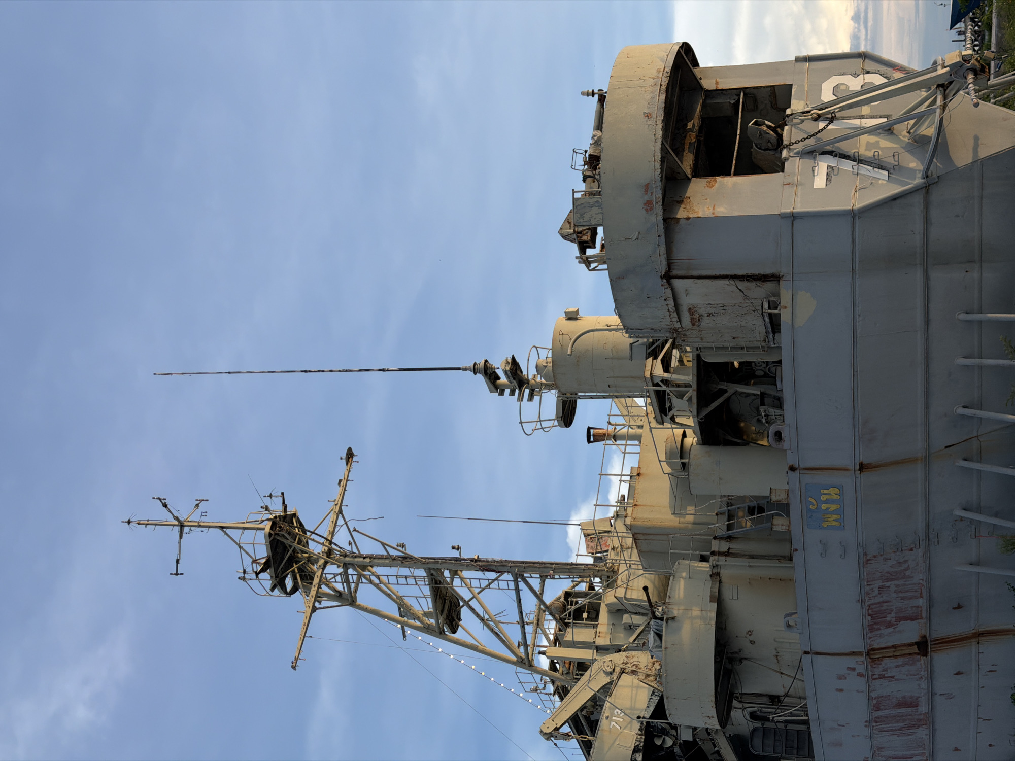 This image shows the stern of a large, gray ship docked in the water. The ship appears to be old and weathered, with rust stains visible on its hull. The ship has a complex superstructure with various antennas and radar equipment. A large number, "713," is painted on the stern. A small Thai inscription, "พงัน," is visible on the side of the hull. The sky above is blue with some scattered clouds. The ship is located in the city of Koh Phangan, Thailand.