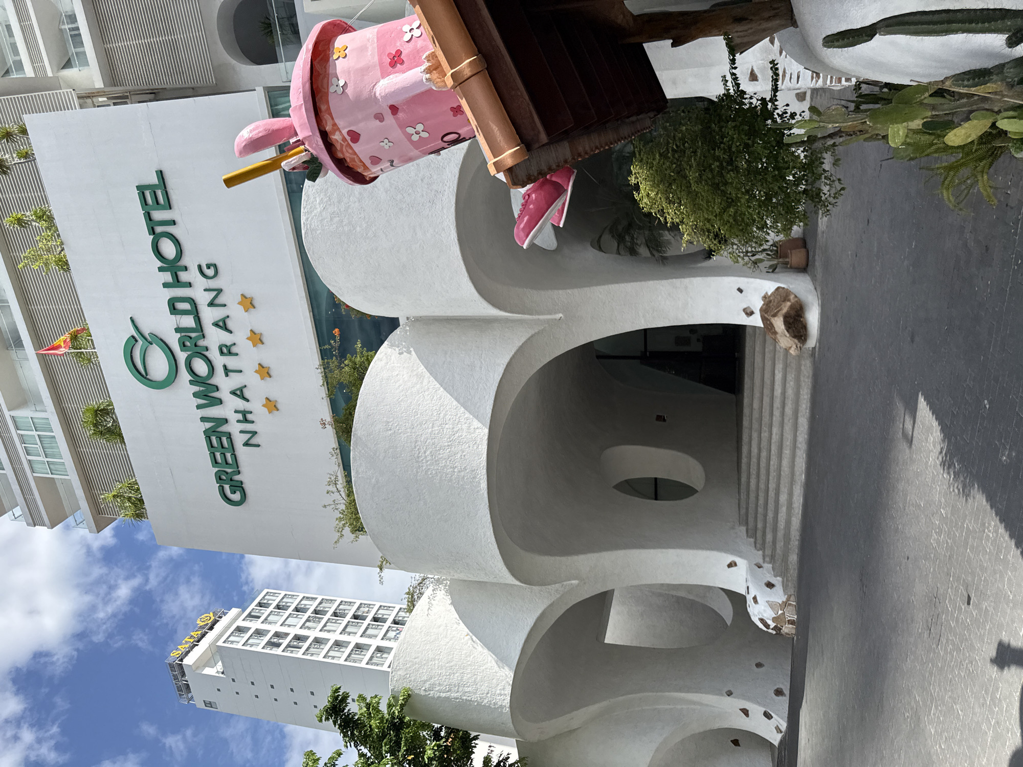 The image shows the exterior of the Green World Hotel in Nha Trang, Vietnam. The hotel has a distinctive white, curvilinear facade with arched entrances. Above the entrance, the hotel's name is displayed in green lettering. To the right of the entrance, a large, whimsical pink sculpture resembling a cup with rabbit ears is prominently featured, with pink shoes dangling from it. In the background, a tall, white building with many windows is visible, possibly another hotel or apartment complex. The sky is blue with some white clouds. The area in front of the hotel appears to be a paved walkway. The scene is brightly lit by daylight, suggesting it is daytime.