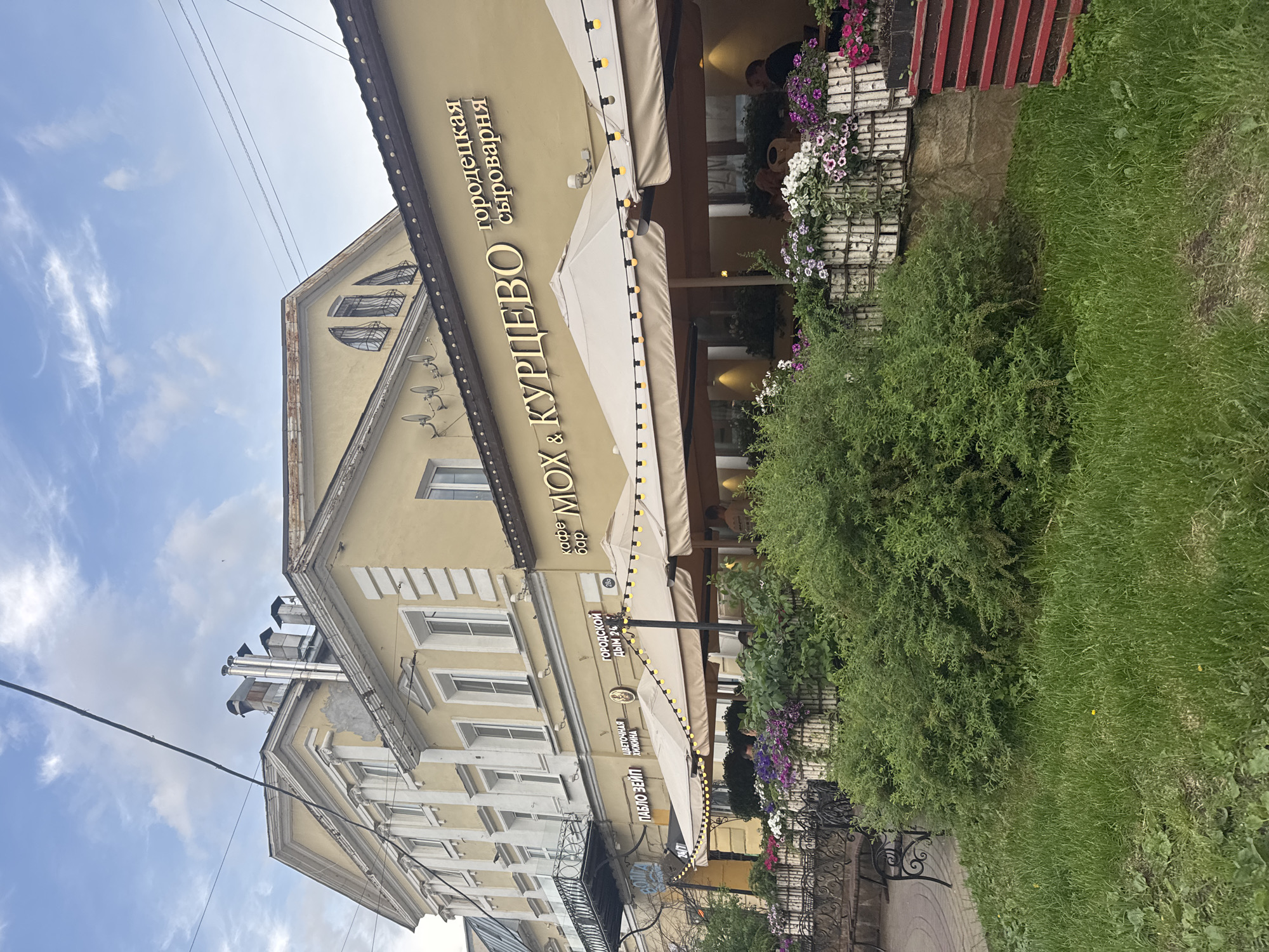 The image shows the building of the restaurant and bar "Mox & Kourtsevo" in Nizhny Novgorod, Russia. The building is a light yellow, multi-storied structure with white window frames. The restaurant has awnings extending over the entrance and outdoor seating area. The restaurant's name is written vertically in gold lettering on the building's facade, next to the text "Городецкая сыроварня". There are outdoor flower boxes along the perimeter. The foreground is green grass, and part of a bush. Overhead electrical wires are visible against the sky. The building's upper floors have a gabled roof with unique arched windows. There are metallic chimneys on top of the roof.