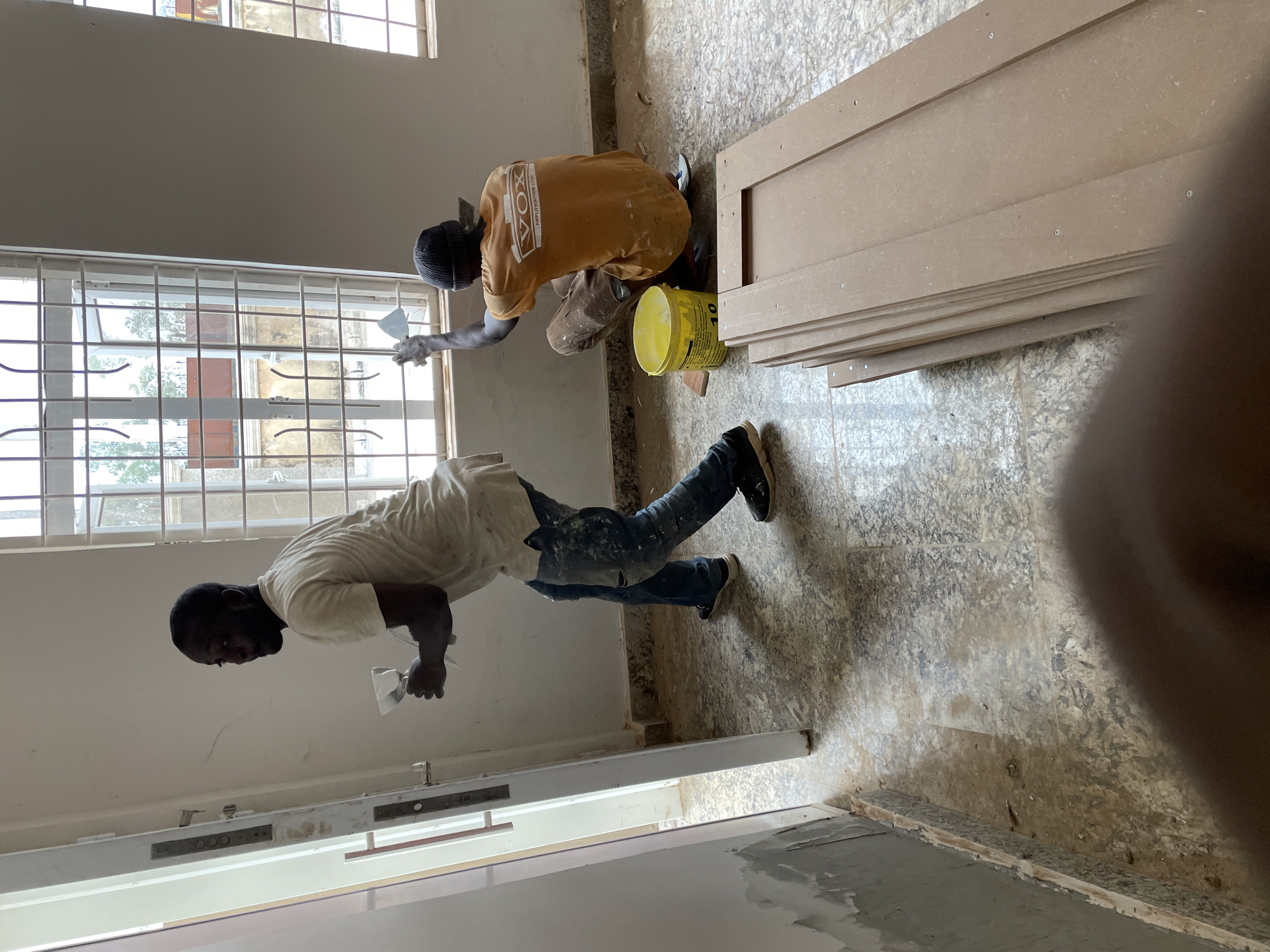 The image depicts an interior construction scene in Jos, Nigeria. Two individuals are engaged in renovation or repair work. One person, wearing a tan t-shirt and dark pants, is seated near a window, applying material, likely filler or caulk, to the window frame. Beside them, a yellow paint bucket with the number "10" is visible. To the right, a stack of what appear to be unfinished wooden panels or doors are propped against the wall. A second individual, wearing a white t-shirt and blue jeans, stands near a doorway, similarly applying material to the wall or door frame. The environment appears to be a room undergoing refurbishment, with unpainted walls and a patterned floor. Natural light is present from the windows.