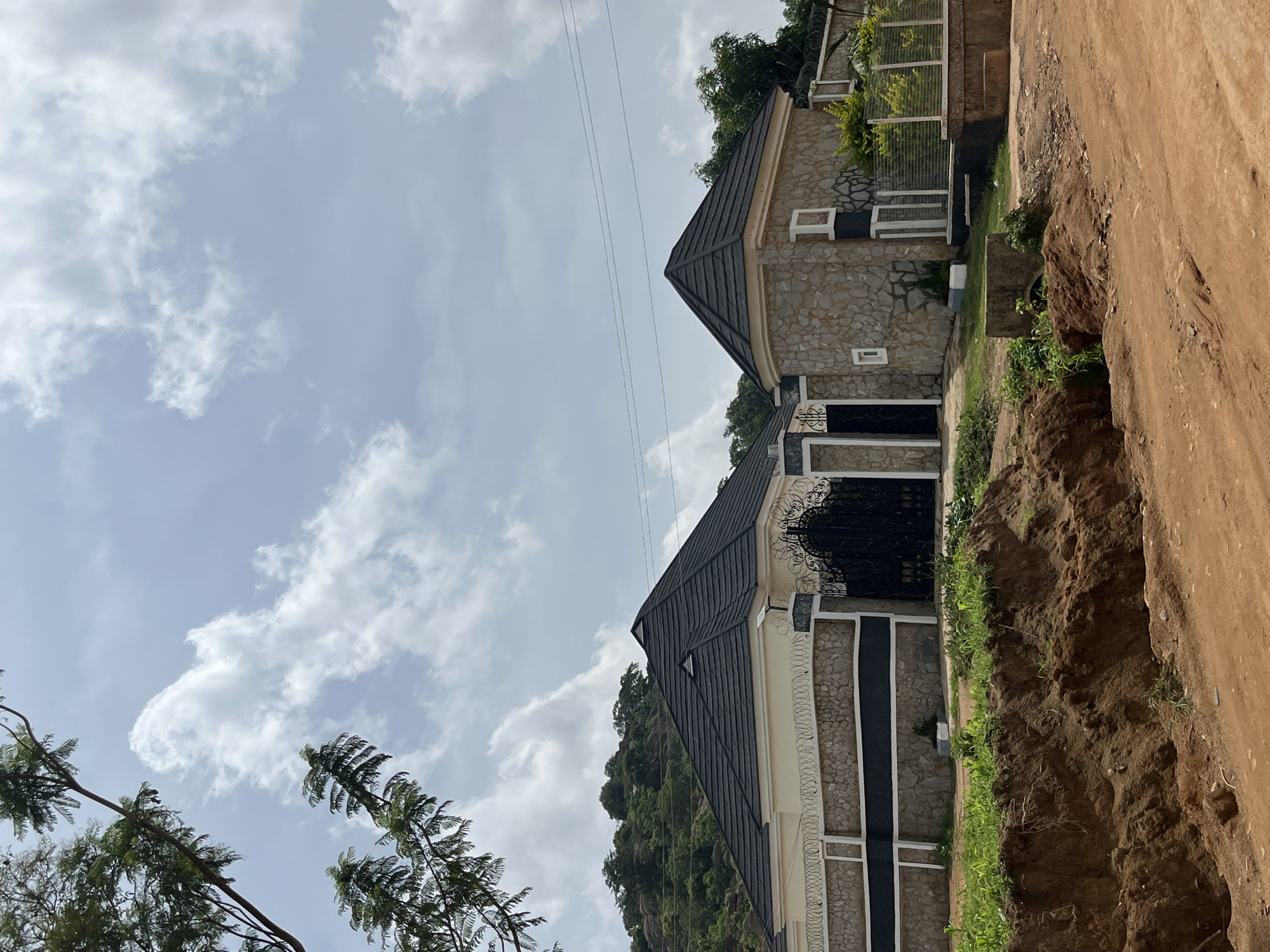 This is a daytime, eye-level, exterior, medium shot of two residential buildings situated on a hillside, possibly in Bukuru, Nigeria, with a dirt road in the foreground. The buildings feature dark, steeply pitched roofs with visible shingles, stone-clad lower walls, and lighter colored upper walls. The larger building on the left has a stone wall topped with barbed wire and a decorative black metal gate. The smaller building on the right is also constructed with stone and has windows with white frames. Lush green vegetation covers the hillside behind the structures, and trees are visible in the upper left quadrant of the image. The sky is partly cloudy with white clouds against a pale blue background. The foreground is dominated by a disturbed dirt embankment and a dirt path, suggesting ongoing construction or landscaping. No people are present in the image.