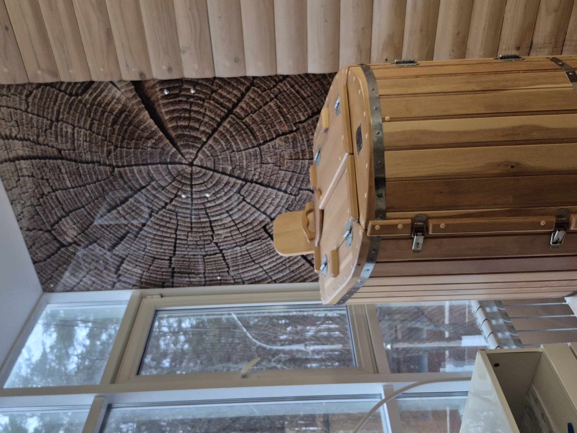 The image shows the interior of a building, potentially located in Chaikovsky, Russia, given the prompt. The ceiling is clad in wooden planks, with a section featuring a large wood grain pattern. A light-colored wooden barrel-like structure with metal bands is suspended near the window. The window reveals an outdoor scene with snow, trees, and possibly buildings in the distance. A section of an indoor structure, probably furniture, appears in the lower right corner of the image. The scene appears to be indoors.