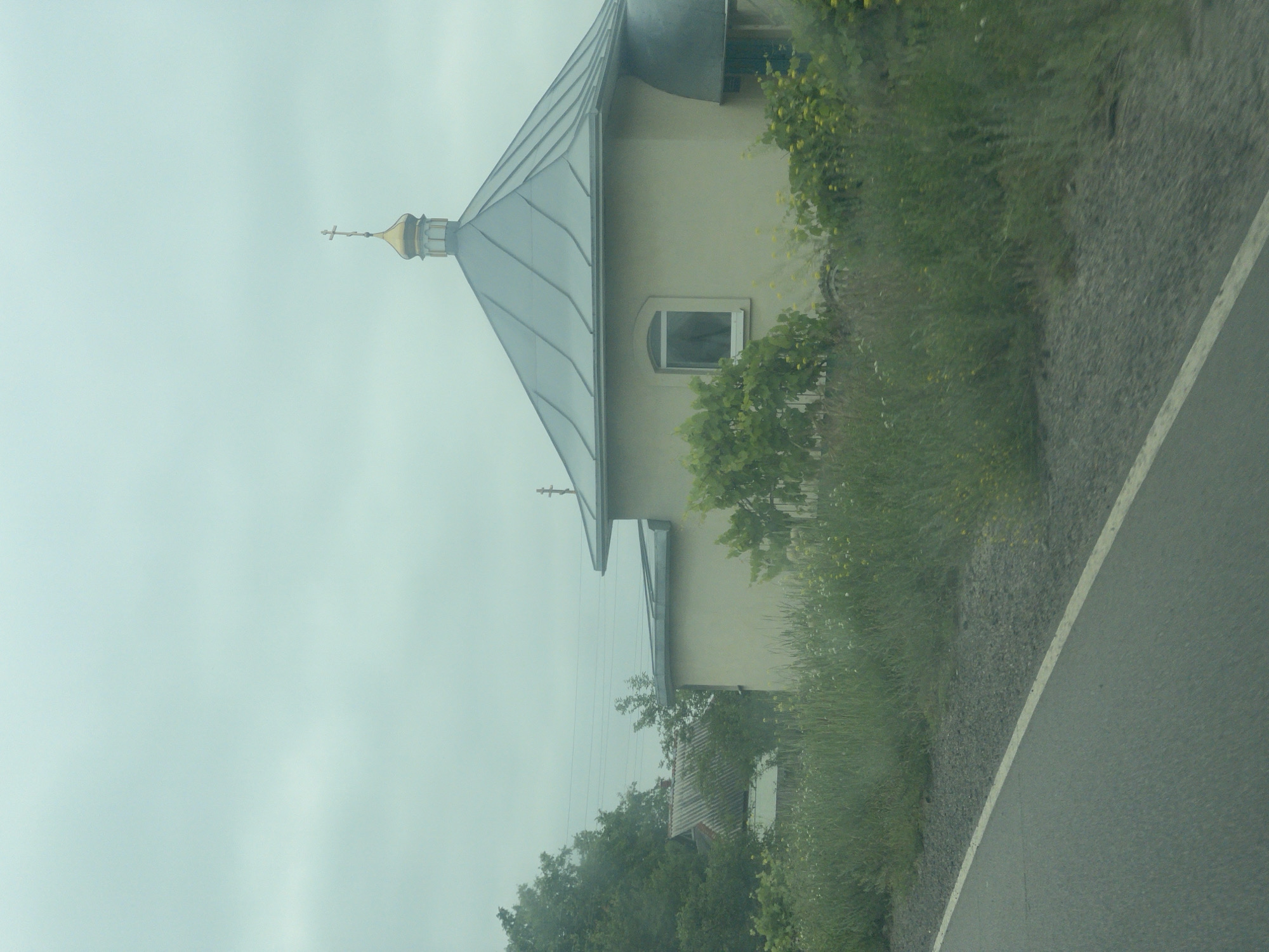 An overcast day is depicted as viewed from a moving vehicle on a road in the village of Kopyli, Ukraine. The scene features a light-colored, one-story building with a distinctive grey, multi-faceted roof topped by a gold dome and cross. A single arched window is visible on the side of the building, partially obscured by green foliage and yellow flowers. To the left, a cluster of trees and another building with a grey, corrugated roof are visible. The road in the foreground is paved and bordered by a gravel shoulder, with tall grass and wildflowers growing along the edge. Power lines are visible in the distance, strung between poles. No people are present in the frame.