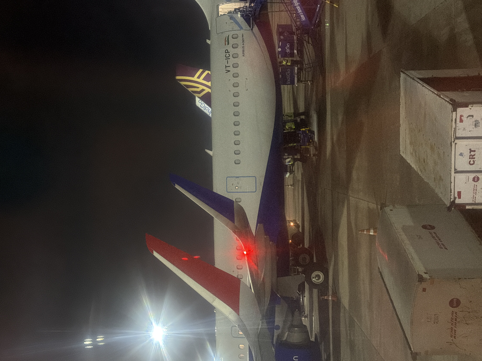 At night, two Indigo Airlines airplanes are parked on the tarmac at Rajiv Gandhi International Airport in Mamidipally, India. The tail of one aircraft, registered as VT-ICP, is visible, displaying the airline's logo. The second aircraft, also an Indigo plane, is behind it, identifiable by its purple and gold tail fin. The planes are illuminated by airport lights, casting long shadows on the concrete surface. Several Indigo airline employees in high-visibility vests are seen near the aircraft, some engaged with baggage containers. A red light is visible on the tail of the first aircraft. In the foreground, two large white cargo containers are parked. The overall scene depicts a typical nighttime operation at an airport.