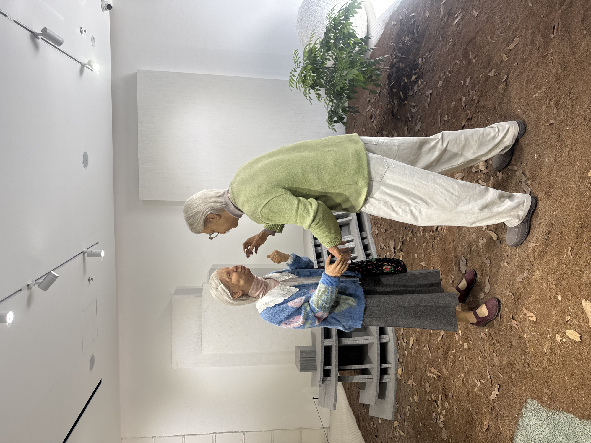 Two lifelike mannequins depicting elderly individuals stand on a reddish-brown, textured ground covered with scattered brown leaves, resembling an outdoor setting within a brightly lit indoor space, possibly an art gallery or museum. The mannequin on the left, facing right, has white hair, wears a blue cardigan with a floral pattern over a pink collared shirt, a gray skirt, and dark shoes. It holds a patterned bag and gestures with its right hand towards the other mannequin. The mannequin on the right, facing left, also has white hair and glasses, wearing a lime green sweater and light-colored trousers with gray shoes. It extends its right hand as if to offer something, while its left hand is partially visible. In the background, there is a grey, multi-tiered structure and a small, leafy green plant. The walls are white, and track lighting is visible on the ceiling, suggesting an interior exhibition space. The overall scene appears to be a diorama or art installation, likely located in Shanghai, China, given the context of such exhibitions in the city.