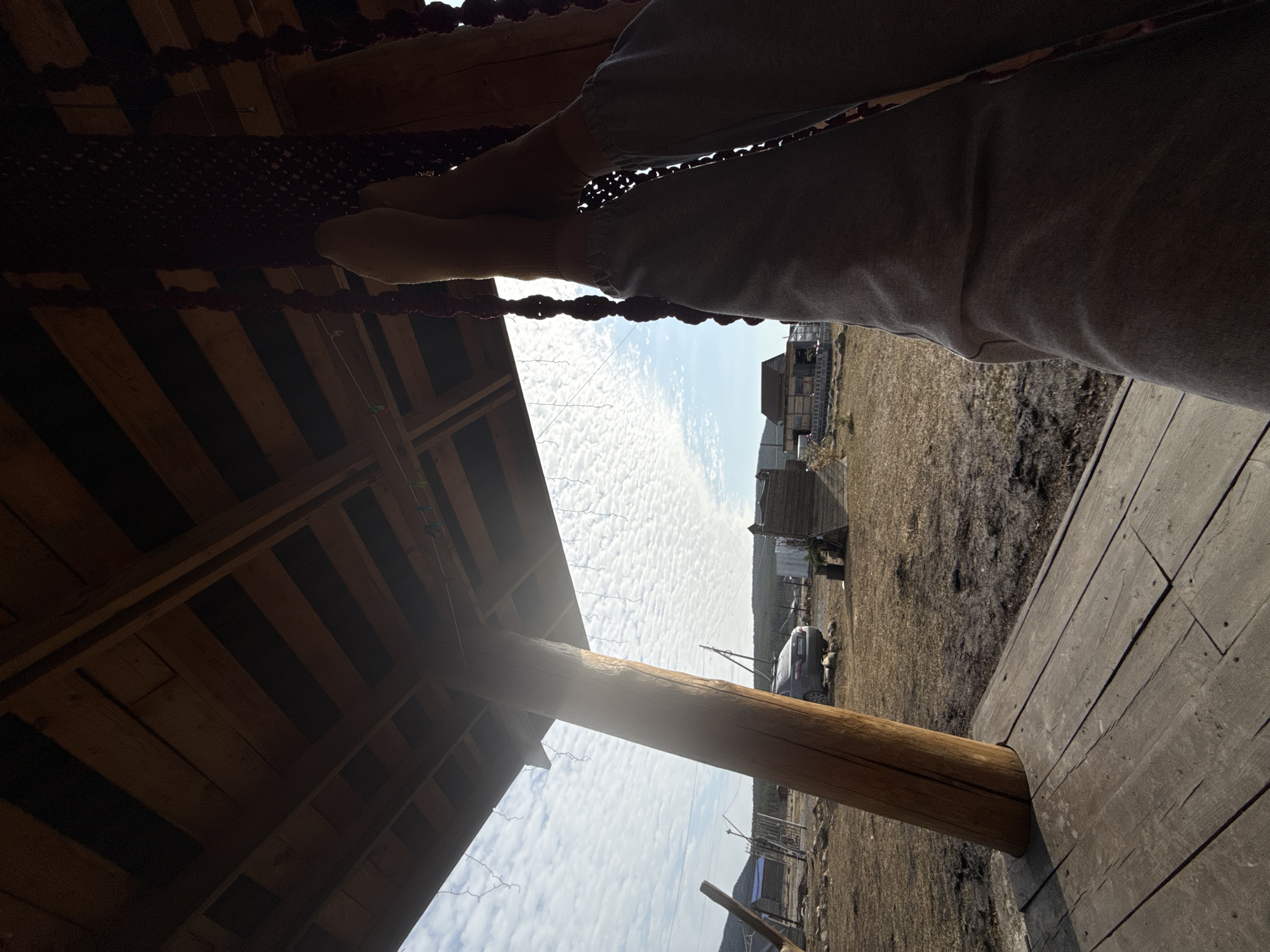 The image depicts a low-angle view from within a wooden structure, possibly a gazebo or shelter, in the rural locale of Tyulyuk, Russia. A person, partially visible and out-of-focus in the upper right, sits within the structure, their clothing light gray. Their bare feet hang in front of them. A burgundy net hangs between the person and the wooden structure.
Outside, the landscape is predominantly dry with patches of grass and bare earth, characteristic of early spring or late autumn. A small cluster of houses, traditional in style, is visible in the distance, along with a gray car parked near a wire fence. The sky is partly cloudy with many small, puffy clouds. The wooden structure is supported by round, wooden poles, with one prominently visible on the left side of the frame. The planks of the structure appear worn and weathered.