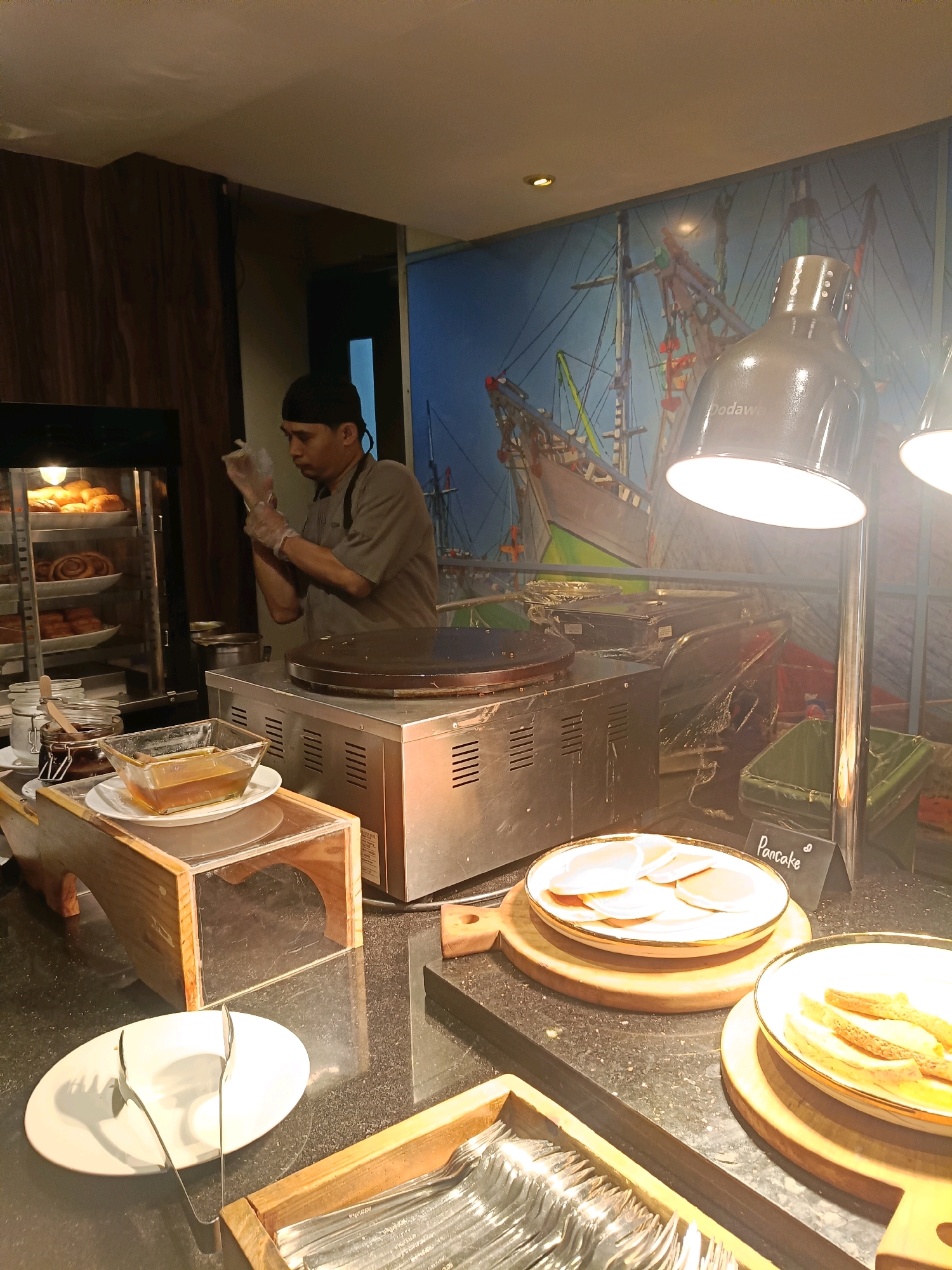 A man wearing a dark cap and a grey uniform, along with gloves, is preparing food at a breakfast buffet. He is standing behind a silver crepe maker, his hands occupied with what appears to be food preparation. To his left, a glass cabinet displays a variety of pastries, including croissants and cinnamon rolls. In the foreground, a small wooden stand holds a glass bowl of what looks like syrup or honey, placed on top of white plates. A stack of white plates with tongs sits on a dark countertop, further to the left. To the right, a plate of pancakes is presented, with a small black sign labeled "Pancake" beside it. Another bowl contains bread slices. The background features a vibrant mural of sailing ships. The overall setting suggests a hotel or restaurant buffet. This scene is likely located in North Jakarta, Indonesia, given the context of the provided image.