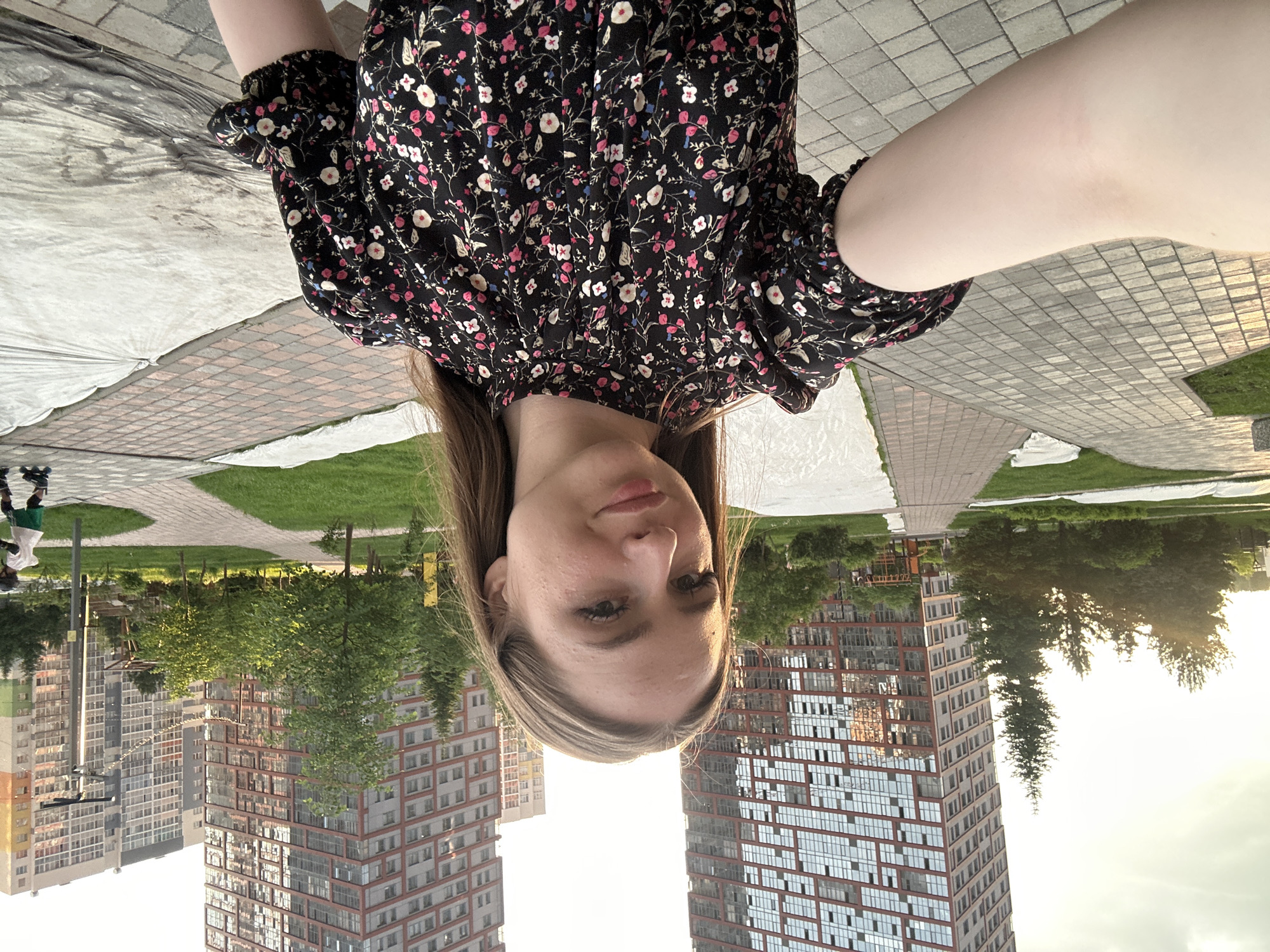 A young woman with long brown hair and light-colored skin is taking a selfie outdoors. She is wearing a black dress with a floral pattern. The background shows modern buildings, trees, and paved pathways. The setting appears to be a city park or residential area. A person on roller skates is visible in the distance on the right side of the frame.
