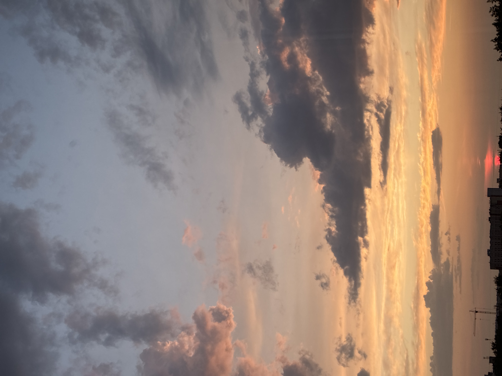 The image depicts a dramatic sunset over a cityscape. The sky is filled with a vibrant palette of oranges, pinks, and blues, with dark gray clouds casting shadows and catching the last rays of sunlight. In the foreground, silhouetted against the colorful sky, are the rooftops of buildings and the skeletal frame of a construction crane. The lower portion of the image reveals the dark outlines of trees and more buildings, suggesting an urban environment. A small, bright red sliver of the sun is visible on the horizon, partially obscured by the buildings. The overall scene is one of natural beauty at the end of the day, set against the backdrop of human habitation and ongoing construction.