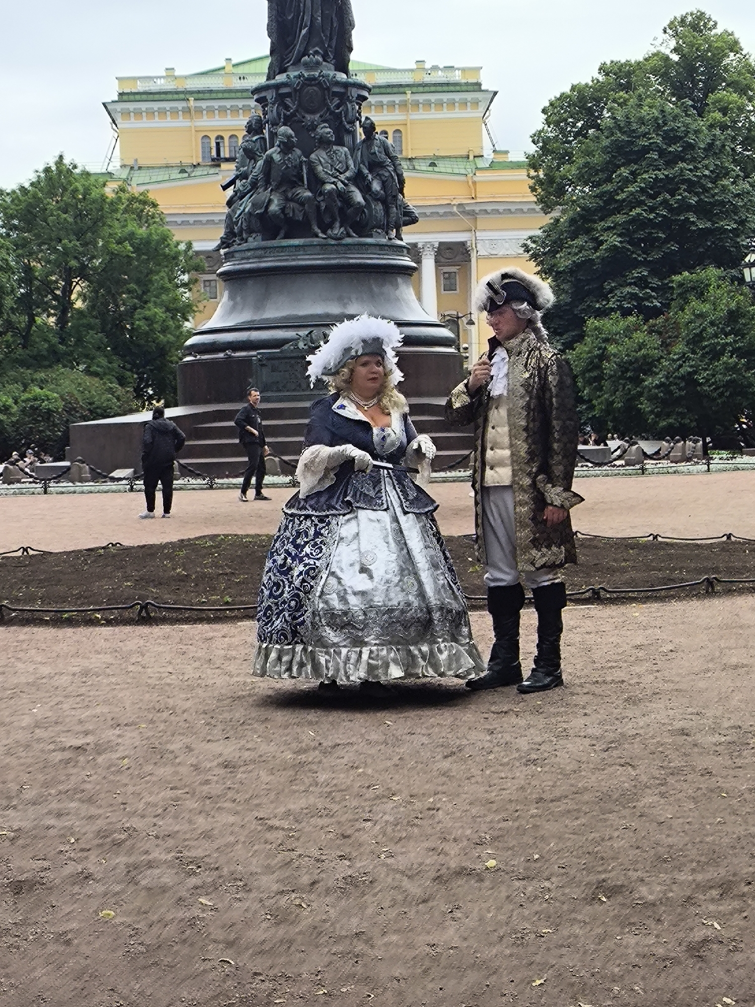 In this image, two individuals dressed in elaborate historical costumes are standing on a dirt path in front of a large monument and a yellow building. The person on the left, appearing to be a woman, is wearing a voluminous silver and blue gown with a white feathered headdress and pearl necklace. The person on the right, seemingly a man, is in a gold embroidered jacket, a white ruffled shirt, grey breeches, and black boots, wearing a powdered wig and goggles.
The monument behind them is dark bronze and features sculpted figures. The yellow building in the background has white architectural details and a green roof, suggesting a grand, possibly historical, location. Trees with lush green foliage frame the scene. The ground is covered in loose dirt and scattered leaves. In the background, other people are visible walking, and there is a dark metal fence separating different areas of the park.
The weather appears overcast, and the lighting is diffused, suggesting it might be daytime, possibly a cool or cloudy day. The overall atmosphere seems to be one of historical reenactment or a themed event, with the two costumed figures being the primary focus. No visible text is present in the image.