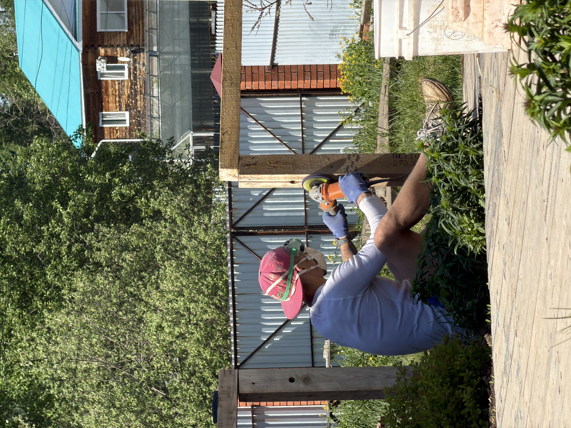 A man wearing blue gloves, a white shirt, a mask, and a red baseball cap is using an orange angle grinder to work on a wooden structure. He is kneeling amidst vegetation. A galvanized metal fence stands in the background, with a brick structure behind it. Further back, a wooden building with a turquoise roof is visible. The photo was taken in Chaikovsky, Russia.
