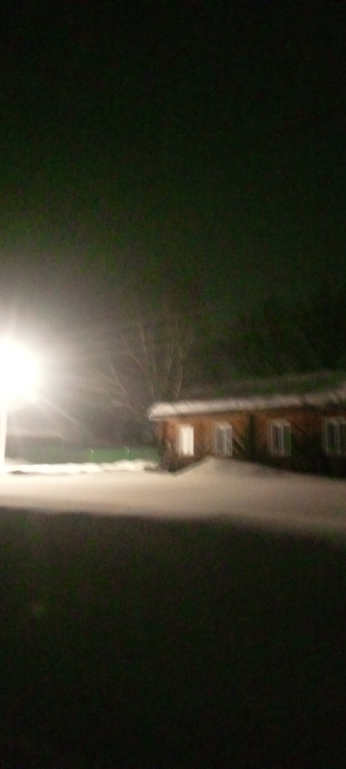 The image depicts a nighttime, snow-covered scene in Красный Маяк, Russia. A single-story, light brown wooden building, consistent with a house or cabin, is situated in the midground, towards the right side of the frame. Its roof is covered with a layer of snow. Multiple rectangular windows are visible along its facade, some appearing to emit internal light. The ground in the foreground and midground is blanketed in snow. A bright artificial light source, possibly a streetlamp or floodlight, is prominently visible on the far left, casting significant glare across the snowy expanse. Bare trees are faintly discernible in the dark background behind the building. No individuals, vehicles, or specific activities are observed.