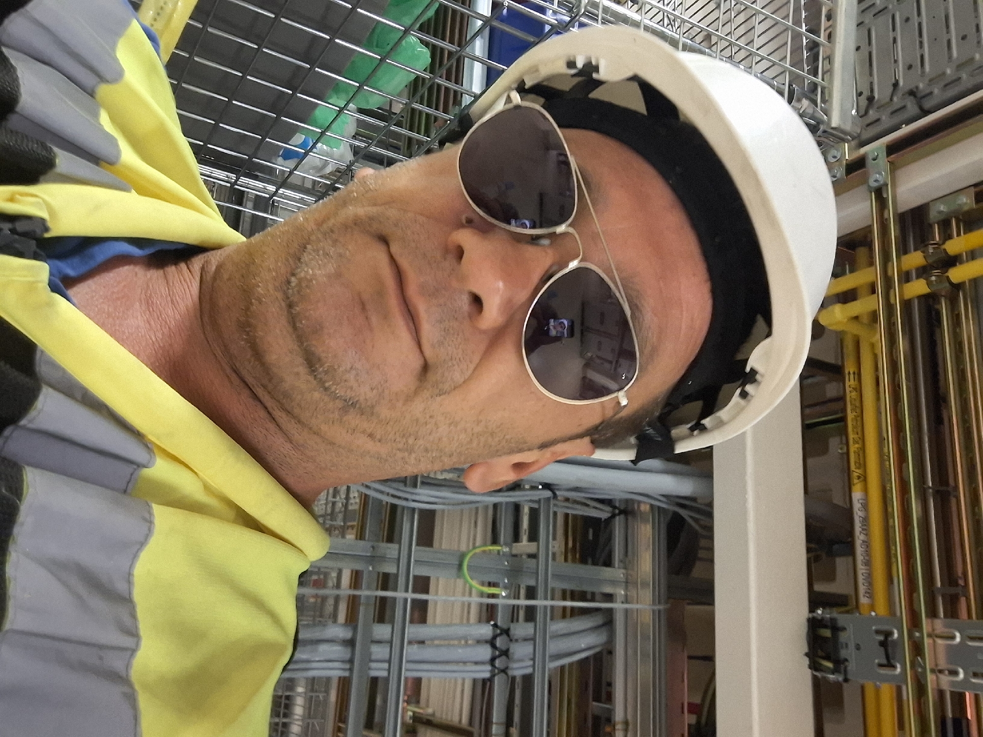 The image is a close-up, selfie-style photograph of a middle-aged man wearing a white hard hat and aviator sunglasses. He is also wearing a high-visibility yellow and gray vest over a blue shirt. The background shows an industrial setting with a complex network of pipes, conduits, and wiring. Specifically, there are yellow pipes with markings, grey conduits, and metal grating. The man's reflection in his sunglasses shows him holding a smartphone and taking the picture. The location is identified as the city of Kiryat Gat, Israel, likely within a construction site or industrial facility.
