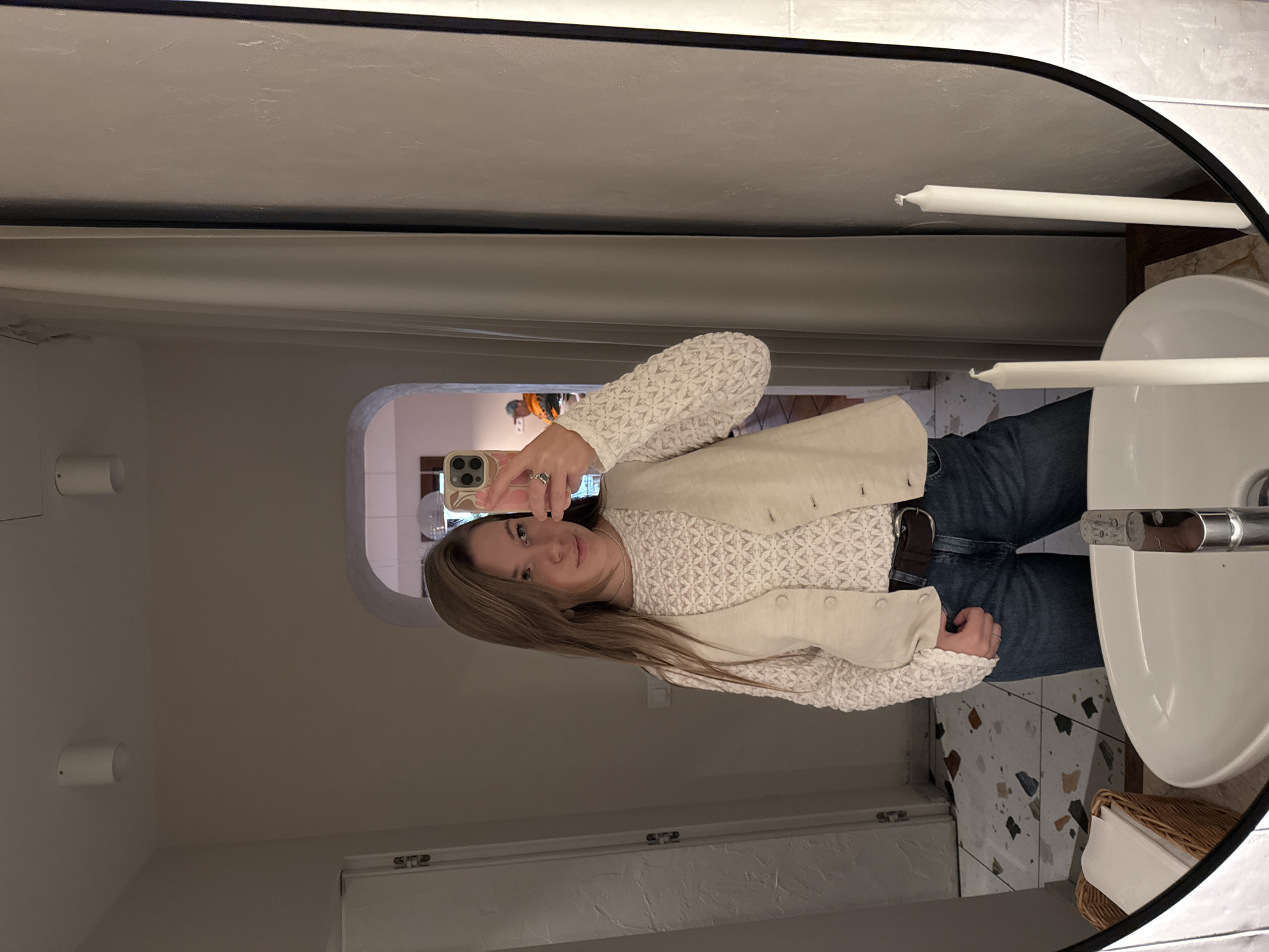 A mirror selfie captures a young woman, likely in her 20s or early 30s, standing in what appears to be a restroom, potentially in Belgrade, Serbia. Her long, straight, light brown hair frames a calm, friendly face as she looks directly into the camera with a slight smile. She holds an iPhone in her right hand, wearing a ring on her ring finger.
Her outfit comprises a cream-colored, long-sleeved top with a distinctive textured, possibly knitted or lace-like, geometric pattern. Over this, she wears a light beige or cream vest adorned with several buttons. She pairs these with blue jeans and a dark brown belt featuring a silver buckle. A simple silver necklace is visible.
The reflection reveals a modern bathroom environment. To the right, a white ceramic sink with a chrome faucet is visible. Behind the sink, two unlit, slender white candles stand upright. The floor features eye-catching terrazzo-style tiles with irregular, multi-colored fragments in shades of brown, black, grey, and beige. A woven wicker basket holding white tissue paper or towels sits on the floor next to the sink.
On the left side of the reflection, two cylindrical white wall-mounted light fixtures are visible. In the background, through an arched doorway reflected in the mirror, another warmly lit interior space with textured walls is glimpsed. The overall lighting is artificial, suggesting an indoor setting without natural light. The walls throughout the visible area have a light, subtle texture. No visible text is discernible in the image.