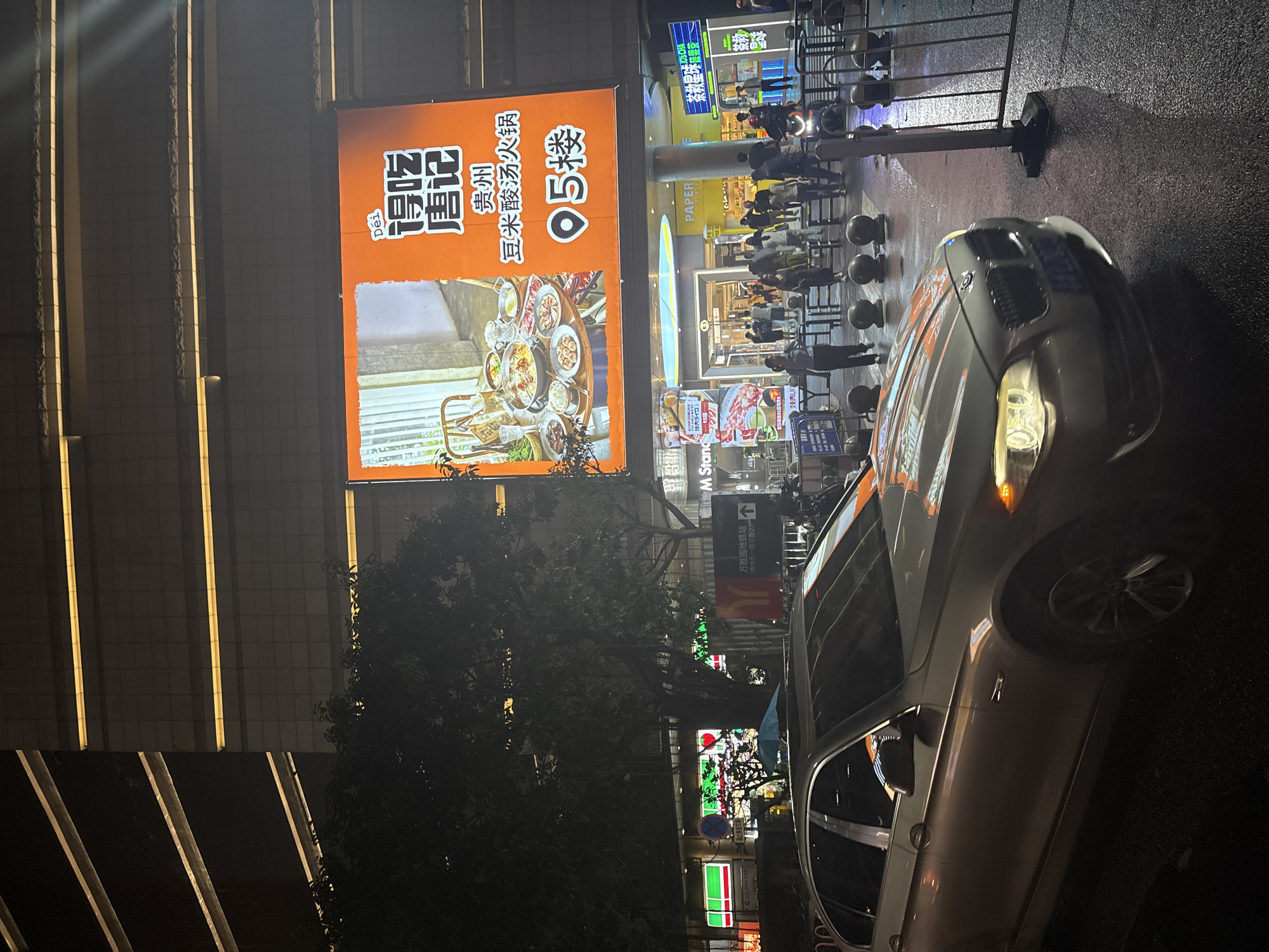 This is a nighttime, outdoor, vertical shot featuring a car parked on a wet street, likely after rain, in front of a brightly lit commercial building. The scene appears to be a busy urban area, possibly a pedestrian street or an area with shops and restaurants.
Dominating the left side of the image is a large, orange billboard advertising a restaurant. The text on the billboard is primarily in Chinese, with "得吃" and "唐记" prominently displayed, suggesting the name of the restaurant. Below that, it reads "贵州 豆米酸汤火锅" which translates to "Guizhou Bean Rice Sour Soup Hot Pot," indicating the type of cuisine offered. A visual of a hot pot meal is also featured on the billboard. To the right of the restaurant name, there's a large "95" which could signify a price or a ranking.
Further back, other signs and shop fronts are visible, including "PAPER", "M Stand", and "茶救星球県茶". Some signs are illuminated with bright colors, contributing to the vibrant, nighttime atmosphere. A sign with Chinese characters and an arrow, "万胜围地铁站 (商场负一层可直达)", indicates that Wanshengwei Subway Station is nearby and accessible from the underground level of the mall.
Several people are milling about in the background, some appearing to be customers or pedestrians. The wet street reflects the artificial lights, adding a glossy sheen to the scene. The overall impression is that of a lively commercial district at night, with food and shopping being the main attractions. The presence of the car suggests it's a street accessible by vehicles. The lighting and the reflections on the wet ground clearly indicate it's dark outside, likely evening or night.