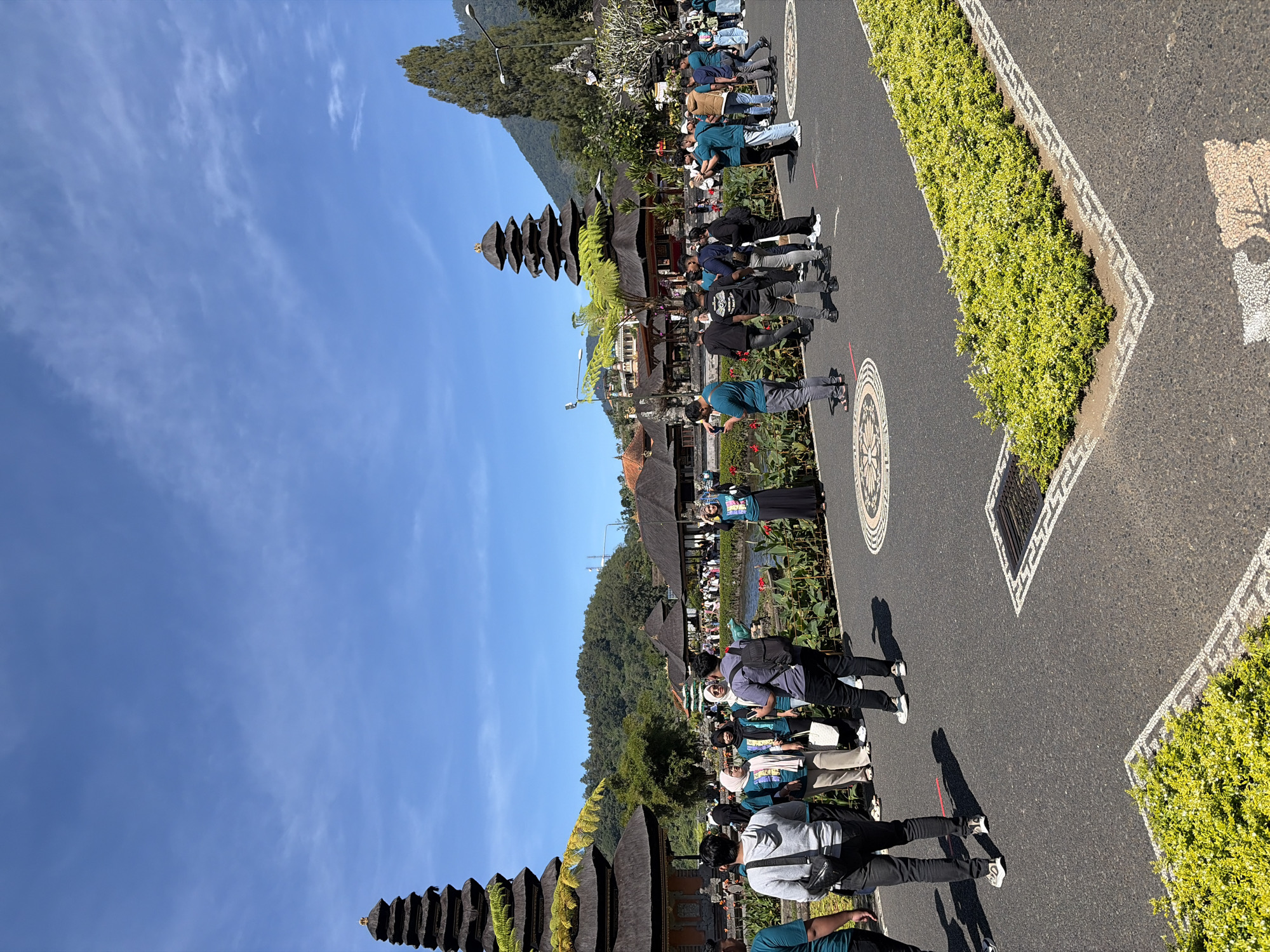 In Baturiti, Indonesia, on a bright, sunny day, a group of tourists stand on a paved pathway in front of a temple complex. The temple features multiple tiered, dark-roofed pagodas, with the most prominent one on the left side of the frame. The architecture suggests a Balinese style. Several individuals are captured mid-action, with some taking photographs, others conversing, and many simply observing the surroundings. Greenery, including manicured bushes and trees, is visible throughout the scene, bordering the pathway and surrounding the water features of the temple. The sky is a clear, vibrant blue with scattered white clouds. The pathway is marked with decorative circular and rectangular inlays.