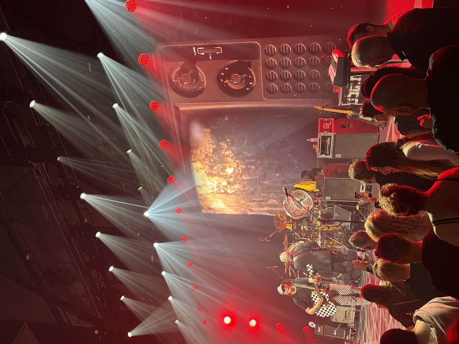 The image depicts an indoor live music concert scene within a venue in Tel-Aviv, Israel. The perspective is from the audience, looking towards the illuminated stage.
On the stage, multiple male musicians are performing. To the left, a man wearing a white cap, black t-shirt, and black-and-white checkered pants plays an acoustic guitar. Next to him, another man in a dark jacket is also playing a guitar, his back partially turned to the audience. Behind them, a drum kit with cymbals is visible, and a drummer is positioned. Additional band members and stage personnel are partially obscured in the background. Musical equipment includes guitar amplifiers, speaker cabinets, microphones, and a keyboard setup on the right side of the stage.
The stage backdrop features a large screen displaying two distinct visuals. The left side shows an abstract, dynamic pattern resembling fiery liquids or flowing lava. The right side displays a graphic representation of a large, vintage-style radio or television set, complete with prominent rotary dials, a toggle switch labeled "LUME," and a grid of speaker perforations.
Overhead, numerous white spotlights project strong, visible beams of light downwards through a hazy atmosphere, creating a dramatic effect. Red stage lights illuminate the stage and foreground, casting a reddish hue over the entire scene. A series of circular red lights are mounted horizontally along the top perimeter of the stage.
In the foreground, a crowd of concert attendees is visible, primarily from behind. Individuals are standing, facing the stage. Several audience members are holding up smartphones, suggesting they are recording video or taking photographs. The overall environment is dark, with strong theatrical lighting focusing on the performers and stage elements.