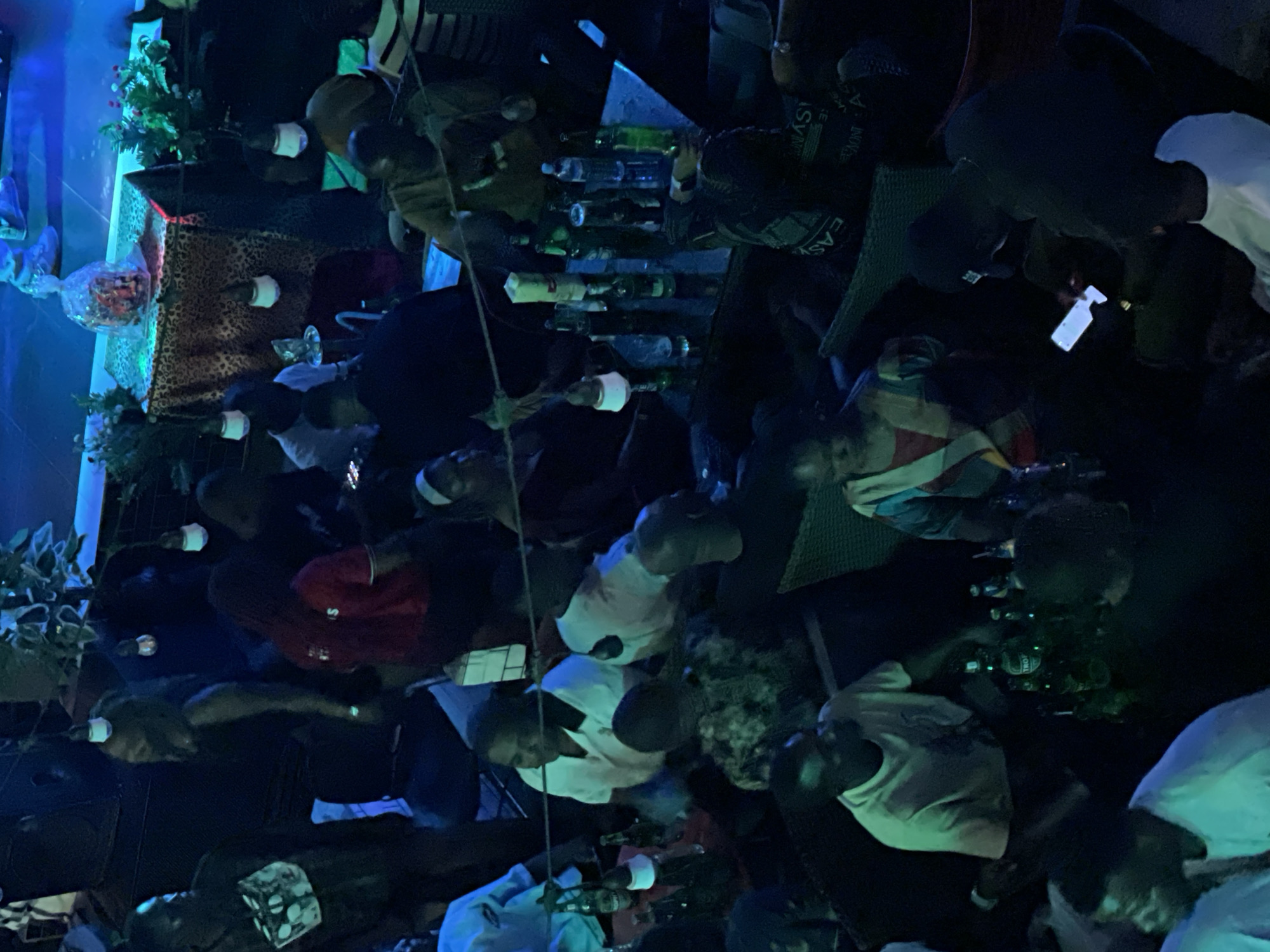 The image depicts an indoor scene, possibly a nightclub or a bar, in the city of Unknown, Unknown. The lighting is dim and primarily blue, suggesting a nighttime atmosphere. Several individuals are present, some seated at tables and others standing.
In the foreground, to the left, a person wearing a white t-shirt is visible. To their right, another person in a white t-shirt is looking away from the camera. A group of people are seated around tables. One person in the center is wearing a light-colored shirt and appears to be looking down. Another person to their right is wearing a colorful striped top. Further to the right, a person in a black cap is holding a phone, which is illuminated.
On the tables, there are several bottles of beverages, some of which appear to be beer bottles. There are also glasses and other items on the tables. In the background, a raised area or stage is visible, with blue lighting. A table covered with a patterned cloth, possibly leopard print, is also present, with a decorative item or basket on top. Plants are also visible in the background. The overall impression is a social gathering in a dimly lit establishment.