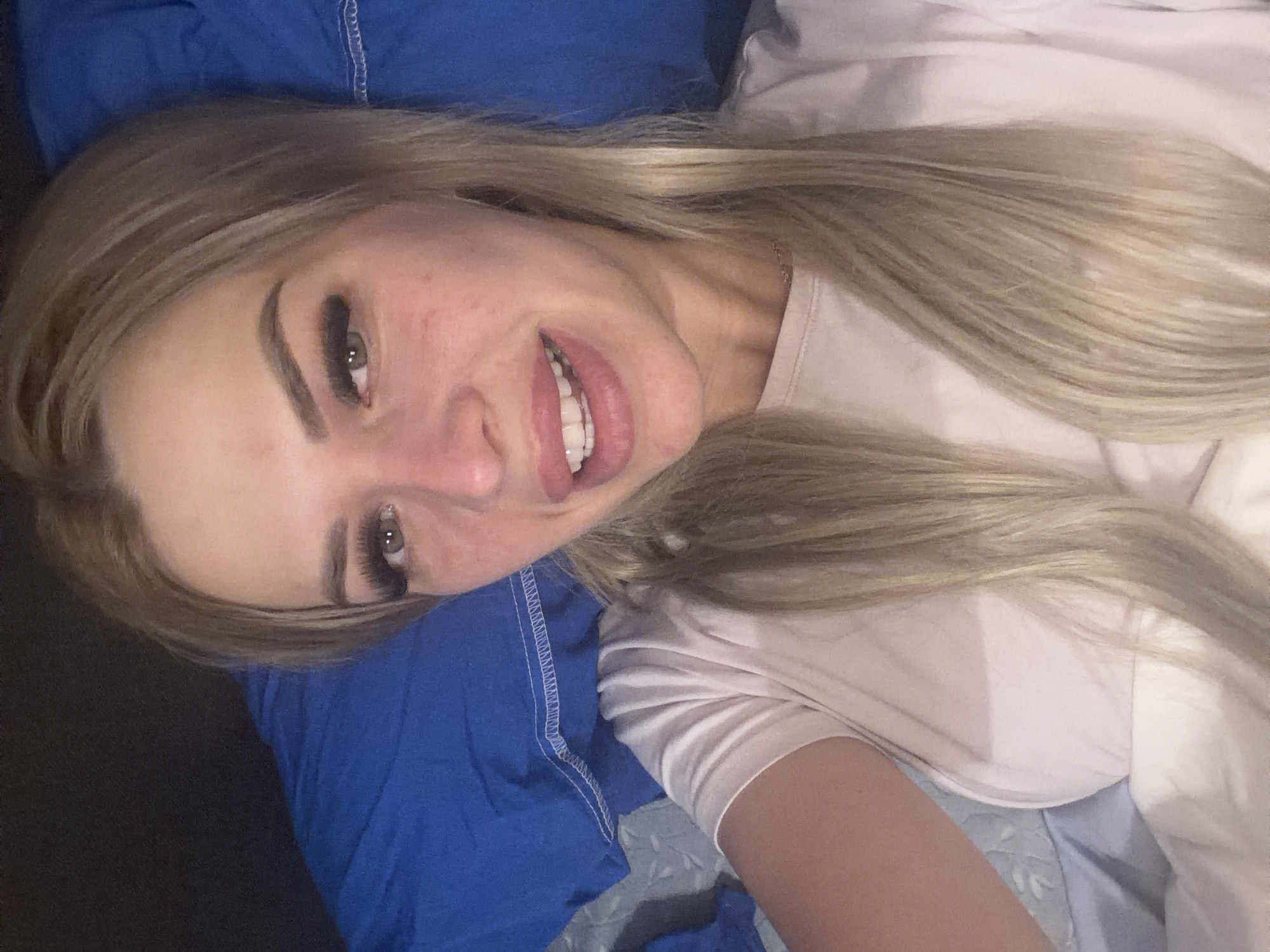 The image is a close-up, selfie-style shot of a young woman lying down, with a blue pillow behind her head and shoulders. She has blonde hair styled in pigtails and is wearing a light pink t-shirt. Her face is looking towards the camera, with her mouth slightly open, revealing her teeth. The lighting suggests an indoor environment, likely a bedroom, possibly at night given the dark background. There are no discernible landmarks or features that would indicate a specific location within Kyiv, Ukraine.