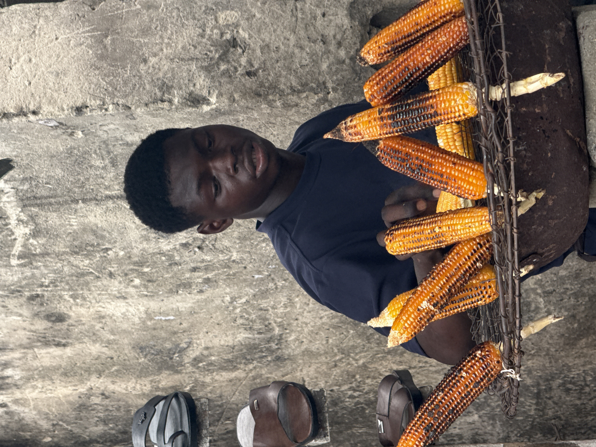 The image, set in Lagos, Nigeria, captures a young Black man engaged in street vending grilled corn. He is depicted from the chest up, wearing a dark blue t-shirt, and looks directly towards the viewer with a neutral expression. His hand is visible near a display of grilled corn.
Numerous ears of corn, exhibiting a mix of golden yellow and charred kernels from grilling or roasting, are arranged on a rustic metal rack. This rack is mounted on or alongside a large, dark, cylindrical object, likely a makeshift grill or cooking apparatus.
The background reveals a textured concrete or paved surface, indicative of an outdoor, urban setting. On the ground in the foreground, a dark backpack and two pairs of sandals (one brown, one grey/black) are visible. The scene portrays a common street food operation during daytime hours. No visible text is present.