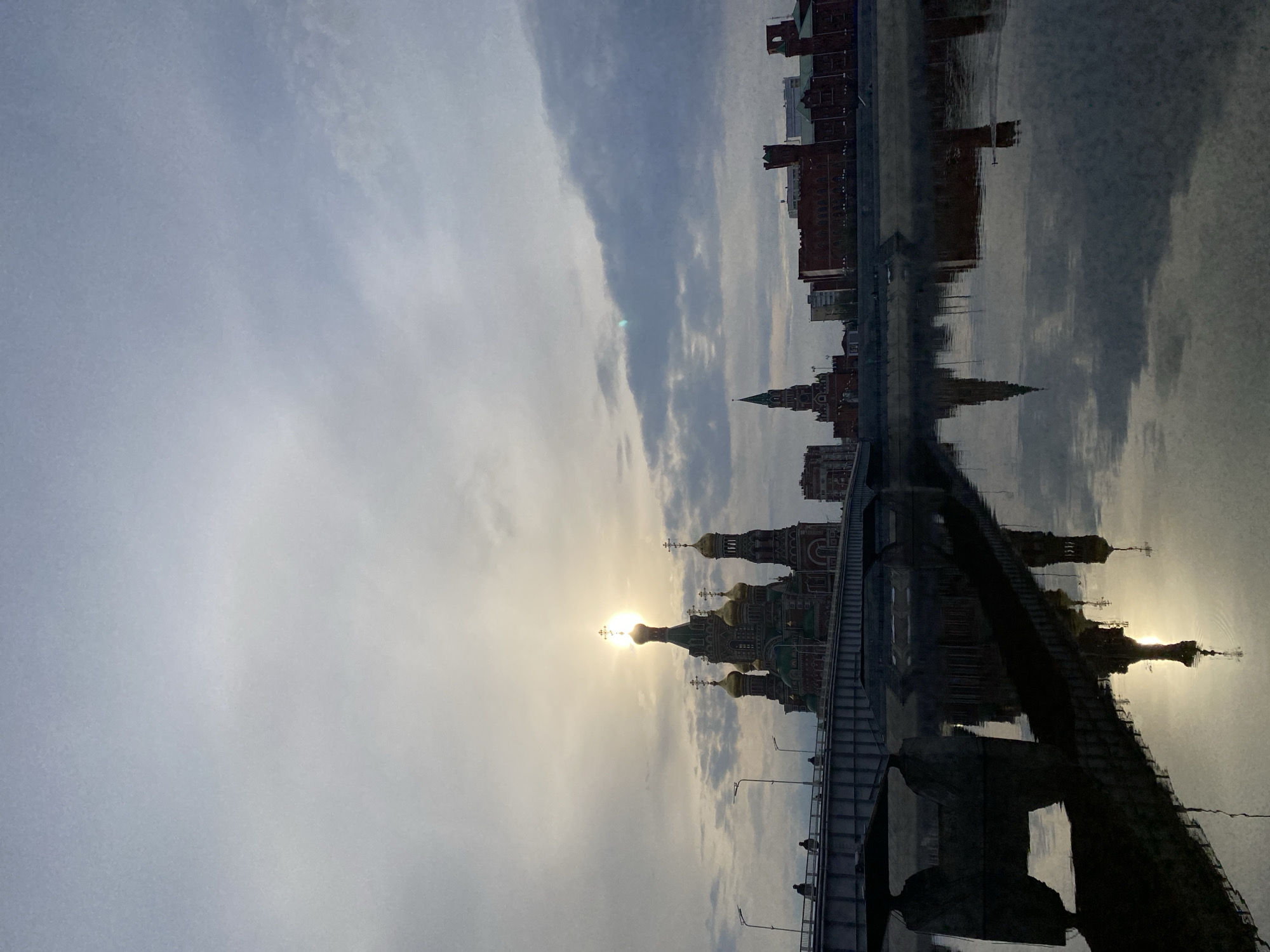 This image presents a serene cityscape of Yoshkar-Ola, Russia, captured at either sunrise or sunset. The scene features a blend of prominent architectural landmarks and modern infrastructure, all reflected in a calm body of water, likely a river or canal.
In the midground, a complex of Russian Orthodox churches with distinctive golden onion domes and crosses stands majestically on the left bank. The sun, low in the sky, is positioned directly behind one of these domes, creating a brilliant halo and a slight lens flare, illuminating the surrounding clouds. The sky is partly cloudy, displaying soft gradients of white and grey.
On the right side, a cluster of city buildings, some featuring red brick facades and one with a notable spire, completes the skyline along the water's edge. A multi-lane bridge, equipped with streetlights and a pedestrian walkway, spans across the water in the foreground and midground. Small, silhouetted figures of people are visible on the bridge's walkway, suggesting movement and daily life.
The tranquil water surface acts as a near-perfect mirror, offering clear reflections of the churches, buildings, parts of the bridge, and the sun's radiant glow. This creates a striking symmetrical composition and enhances the overall peaceful ambiance. No text is visible in the image.