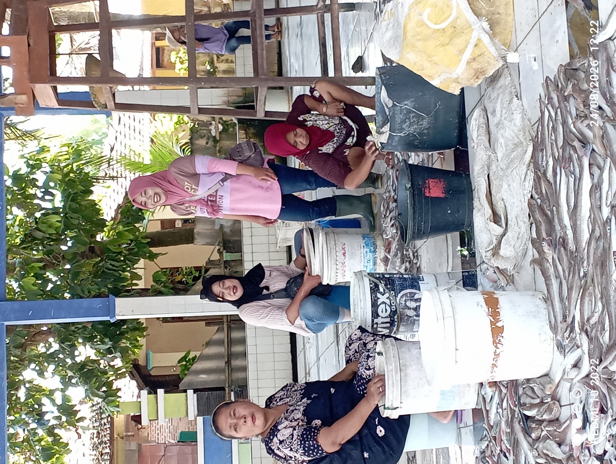 The image depicts an outdoor market scene in Karangsari, Indonesia, with four women and a child visible. The primary subjects are two women in the foreground. The woman on the left, wearing a dark blue patterned dress and a headscarf, is seated and looking to the left, with her hands resting on a white bucket. To her right, a woman in a pink and brown patterned shirt and blue jeans is crouched, wearing a black headscarf and looking at the camera. Behind her, another woman in a pink hijab and a light pink and brown long-sleeved shirt is laughing and looking to her left. In the background, a child in a purple shirt and blue jeans is sitting on a wooden structure. The ground is covered with numerous small fish, buckets, and tarps. The environment suggests a market or processing area, likely for fish, with elements like tiled flooring and a covered structure overhead.