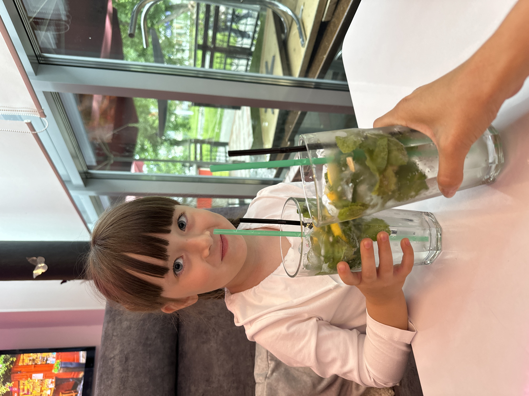 A young girl with brown hair and blue eyes is featured in this vertically oriented, close-up photograph. She is wearing a white long-sleeved shirt and is looking directly at the camera with a neutral expression. The girl is positioned in the foreground, with her face and upper body visible.
In front of her are two glasses of a beverage, likely a mojito, filled with ice, mint leaves, and lemon slices. Two straws, one green and one black, extend from the glasses. A hand, presumably belonging to an adult, is holding one of the glasses.
The setting appears to be an indoor space with large windows. Through the windows, a glimpse of an outdoor scene can be seen, with trees and greenery suggesting a daytime setting, possibly late spring or summer. The lighting is bright, indicating daylight. Part of a television screen showing a colorful image is visible in the lower left corner of the frame. The overall atmosphere is casual and relaxed, possibly a cafe or restaurant. No text is visible in the image. The location context is provided as Chaikovsky, Russia.