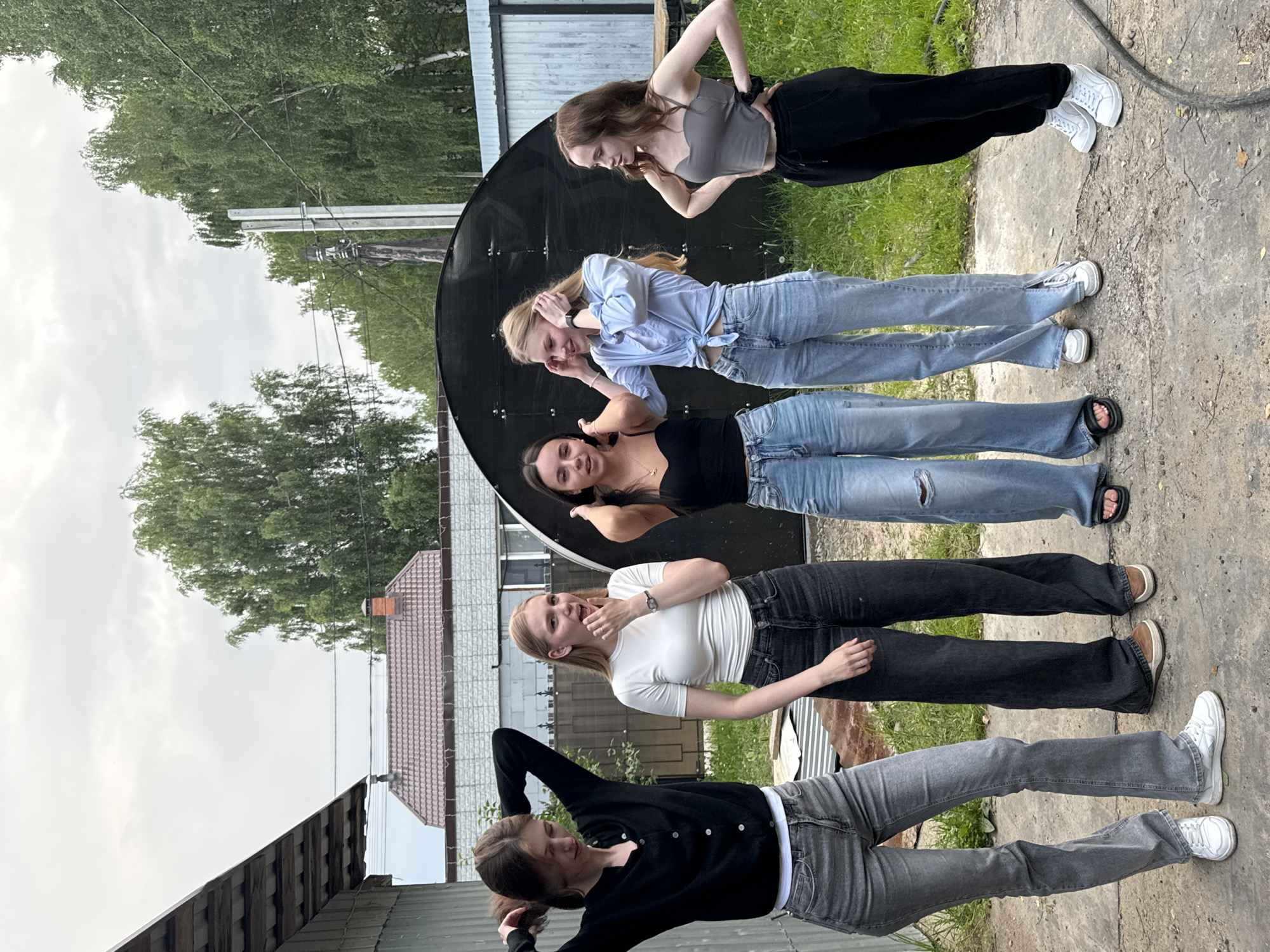 The image shows a group of five young women in casual clothing posing outdoors. They appear to be in a rural or semi-rural setting, with buildings, trees, and a cloudy sky visible in the background. The women are arranged in a line, with some standing and others leaning. They are wearing jeans and various tops, including t-shirts and a button-down shirt. One woman is wearing a black cardigan and grey jeans, another a white t-shirt and black jeans, a third a blue button-down shirt and jeans, a fourth a grey tank top and black pants, and the fifth is mostly out of frame but appears to be wearing a black top and black pants. A large, dark, circular object, possibly a satellite dish or a decorative element, is positioned behind some of the women. The lighting suggests it is daytime, possibly overcast. The overall mood seems relaxed and playful. There is no visible text in the image. The context provided indicates the location is Perm, Russia.