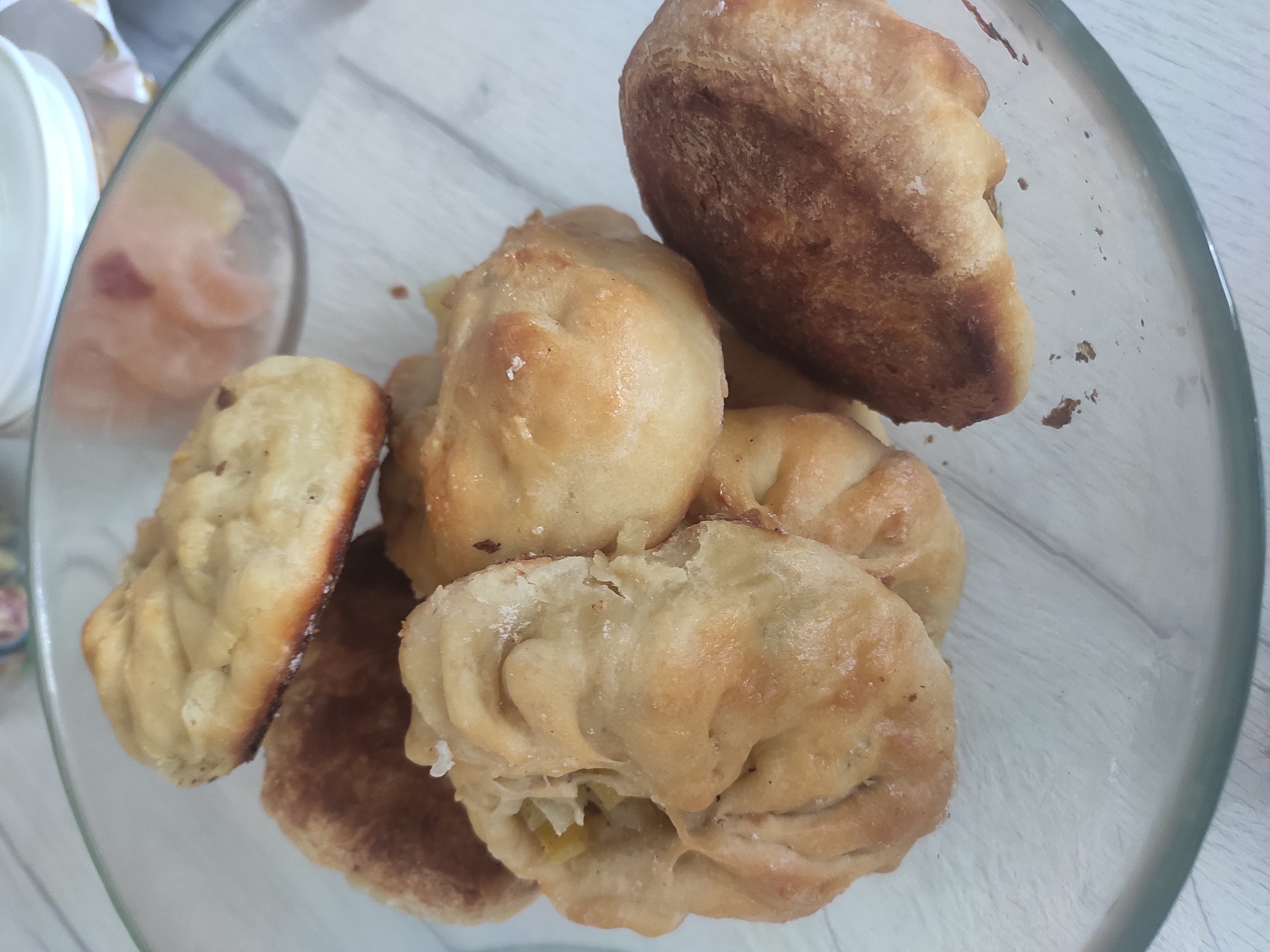 The image shows a clear glass bowl filled with baked goods, possibly buns or pastries. The baked goods have a golden-brown color and appear to be slightly greasy. The bowl sits on a light-colored, possibly wooden, surface. There is another container visible in the background, partially filled with something that appears orange and white. The location is mentioned as Sterlitamak, Russia, but there is no visual information in the image to confirm this.