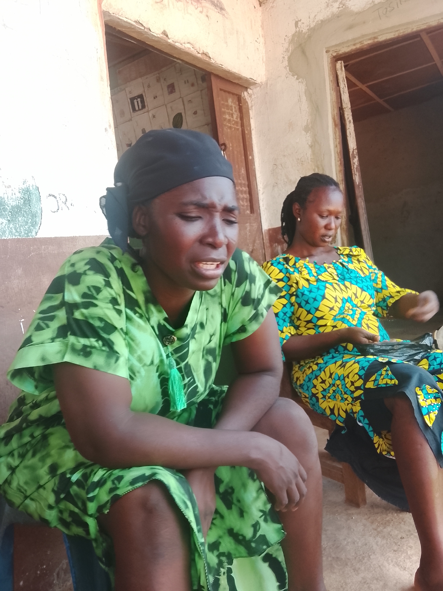 In Akwanga, Nigeria, two women are seated outside a weathered building. The woman on the left, wearing a black headscarf and a green tie-dye dress, appears distressed, with her eyes closed and mouth open in what could be a cry or expression of pain. Her left arm rests on her lap, and her right arm is bent at the elbow. To her right, another woman is seated, wearing a yellow and blue patterned dress. She is looking down and holding a dark object, possibly a bag, in her lap with both hands. The building behind them has a rough, textured exterior with some graffiti. The doorway is open, revealing wooden beams and a glimpse of the interior. The ground appears to be concrete.