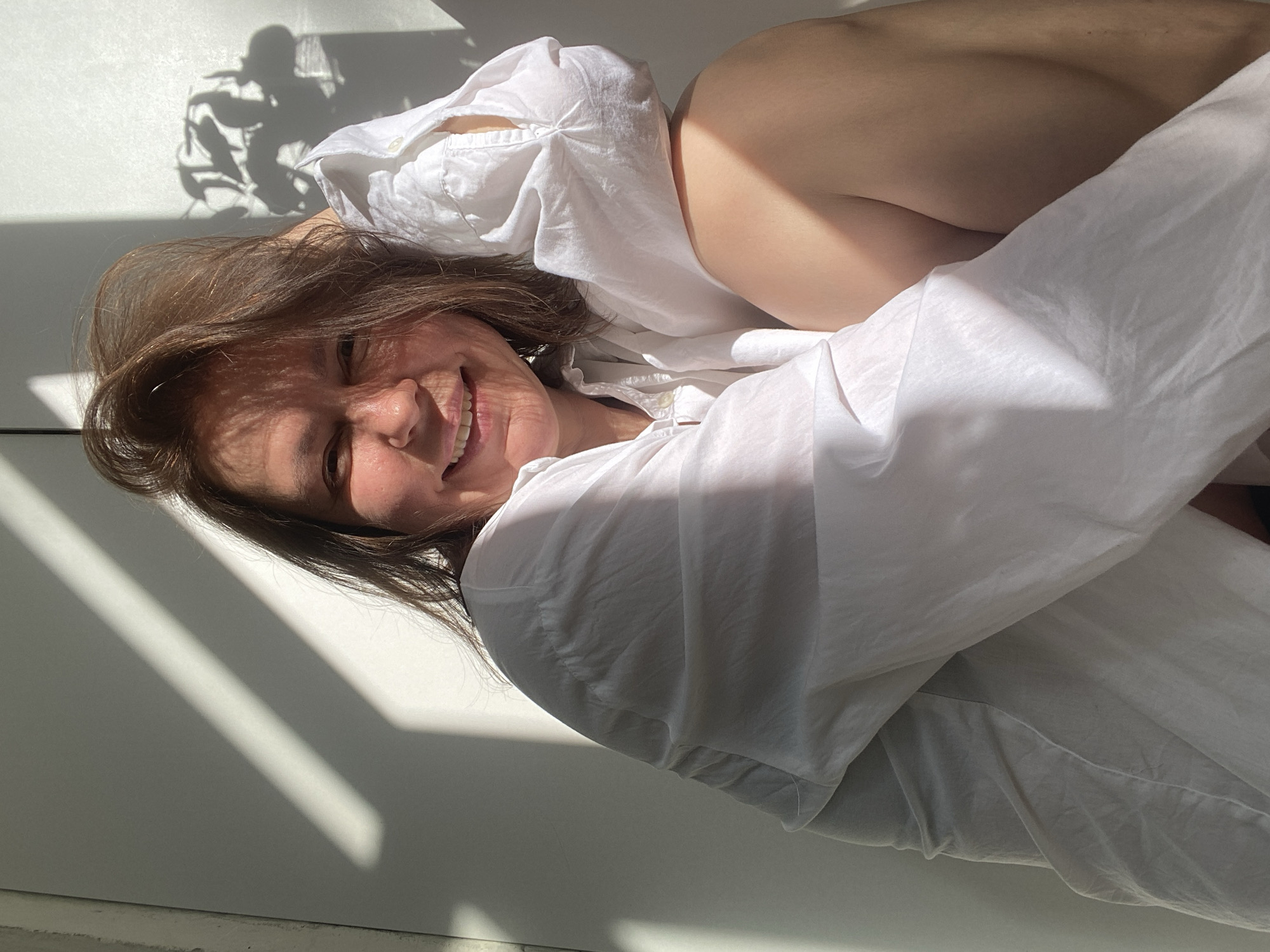 A woman with short brown hair is lying down, smiling, and wearing a white shirt. Sunlight is streaming into the room, casting shadows on the wall and the woman's face. The location is in Chaikovsky, Russia, indicated by metadata not visible in the provided image.