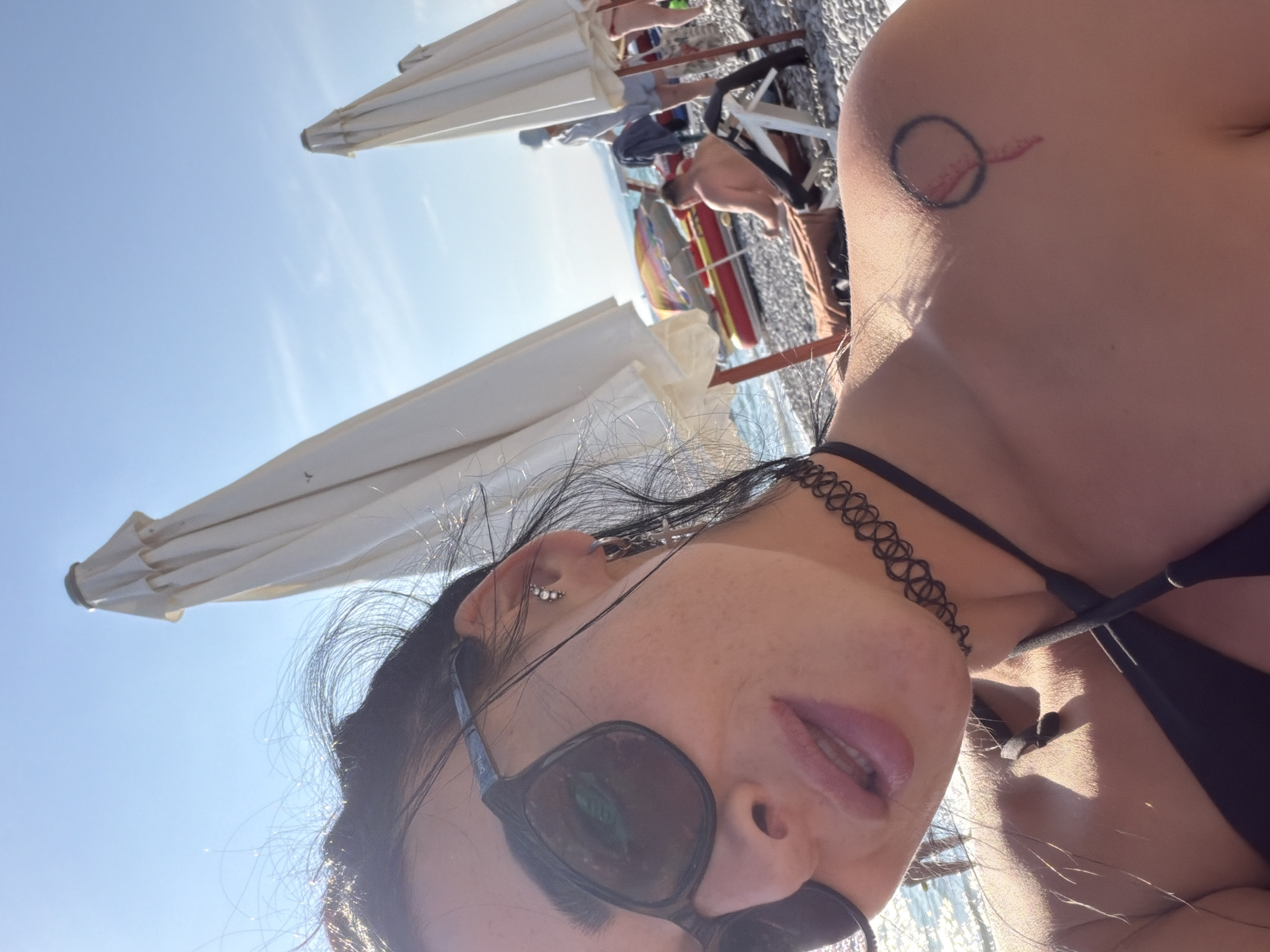 A selfie taken at the beach in Shepsi, Russia, features a woman in the foreground wearing sunglasses and a black bikini top. She has a black choker necklace and a tattoo on her left shoulder. In the background, several closed beach umbrellas, beach chairs, and other beachgoers are visible. The sea and sky are also present.
