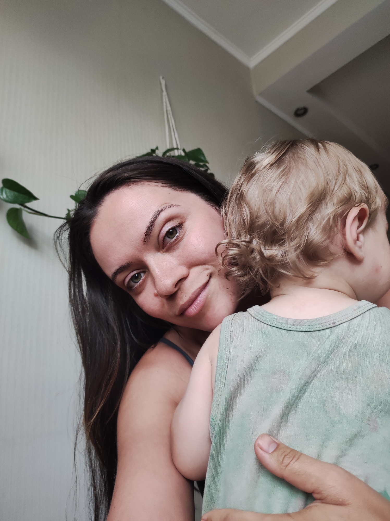 A woman with long, dark hair is holding a toddler who is facing away from the camera. The woman is looking at the camera with a slight smile. The toddler is wearing a light green tank top. The background appears to be an indoor setting with a plain wall and a plant hanging from the ceiling. There is no identifiable signage or architectural features that would indicate a specific location within Chaikovsky, Russia. The image is taken from a close-up perspective.