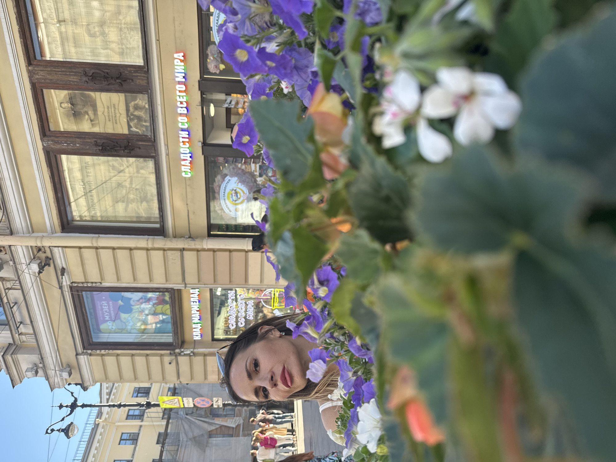 A woman, partially obscured by blooming purple and white flowers, looks towards the camera. She has dark hair and is wearing sunglasses perched on her head. In the background, a building with ornate architectural details features several large windows. One window displays a colorful stained-glass image with cartoon characters, and signage indicates "МУЗЕЙ ПАНОМАЛИЙ ТЕЛА" (Museum of Body Anomalies). Another sign reads "СЛАДОСТИ СО ВСЕГО МИРА" (Sweets from all over the world), suggesting a street scene with shops. The lighting suggests it is daytime, likely a sunny afternoon given the clear sky visible in a lower portion of the image. The overall impression is a casual snapshot taken outdoors, possibly during a stroll through a city.
