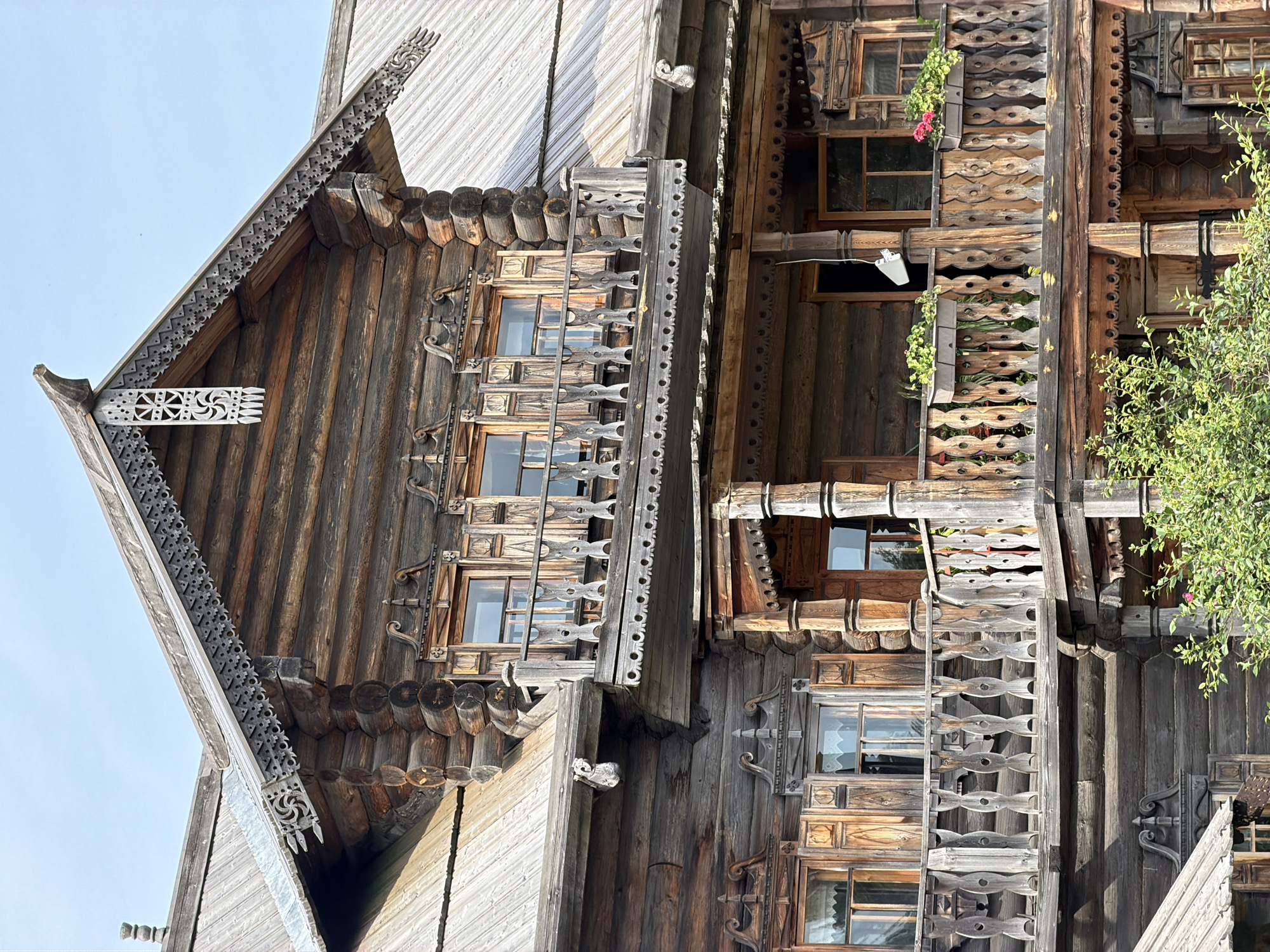 The image depicts a detailed view of a traditional wooden log house with ornate carvings, likely in Imeni Sverdlova, Russia. The architecture features multiple levels, including balconies with intricate wooden railings and decorative eaves adorned with carved patterns. The house is constructed from thick, horizontal logs. Several windows are visible, some with wooden shutters. On the upper balcony, there are window boxes with green plants and some red flowers. The overall scene is one of rustic, traditional craftsmanship in a natural outdoor setting, with a clear blue sky in the background. There are no people or animals visible in the image. The focus is entirely on the architectural details of the building.
