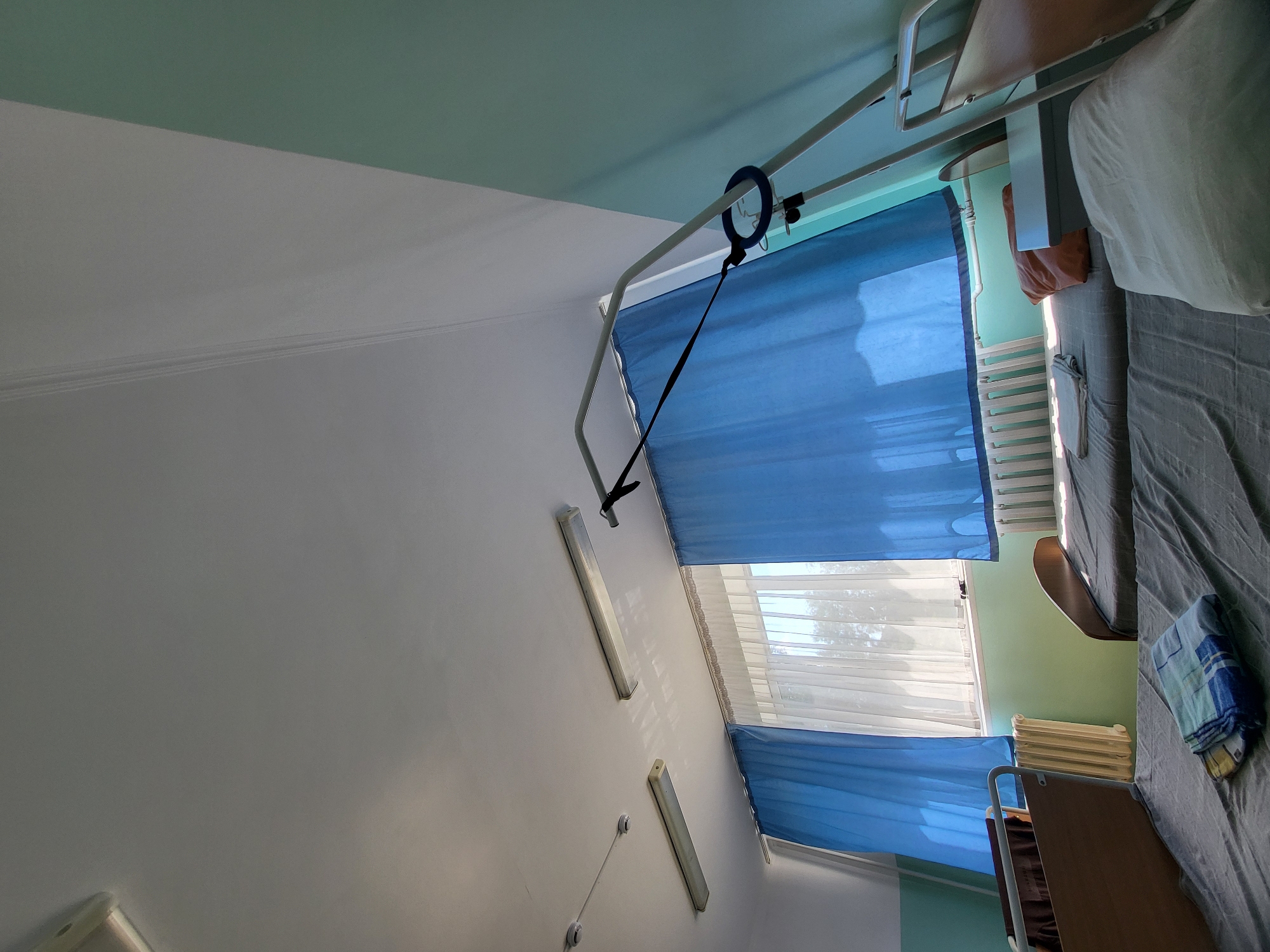 The image depicts a room that appears to be a hospital ward or a similar healthcare facility. The room contains two beds with grey bedding. On the bed in the foreground, there is a folded blue and white striped towel. The bed on the right has white bedding and an orange pillow. The room is lit by natural light coming through a large window covered by sheer white curtains and blue drapes. A radiator is visible beneath the window. Mounted on the ceiling are two long fluorescent light fixtures. A medical bed frame with a sling is positioned over one of the beds. The walls are painted in a light green color. The image does not provide any specific visual cues to determine its exact location within Petropavl, Kazakhstan.