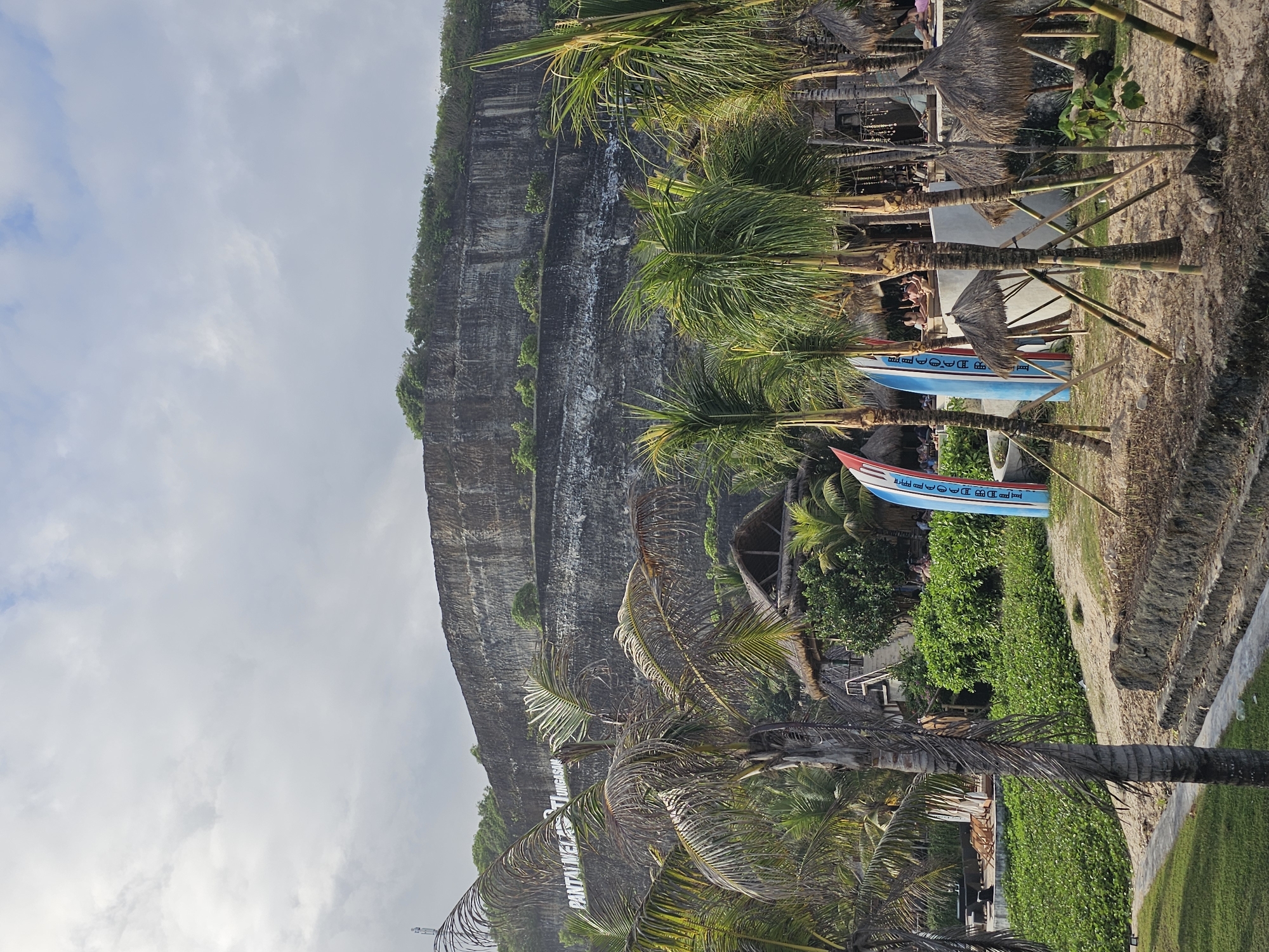 The image depicts a coastal scene, likely at Pantai Melasti, in Kuta Selatan, Indonesia. A tall, rocky cliff dominates the left side of the frame, with vegetation clinging to its surface. The sky is overcast with scattered clouds. In the foreground, there are several palm trees, some of which are partially blocking the view. Two fishing boats, painted in blue and red, rest on the sandy ground. Thatched-roof structures can be seen amidst the greenery and trees. The vegetation appears lush and tropical. In the distance is a sign that reads "PANTAI MELASTI".