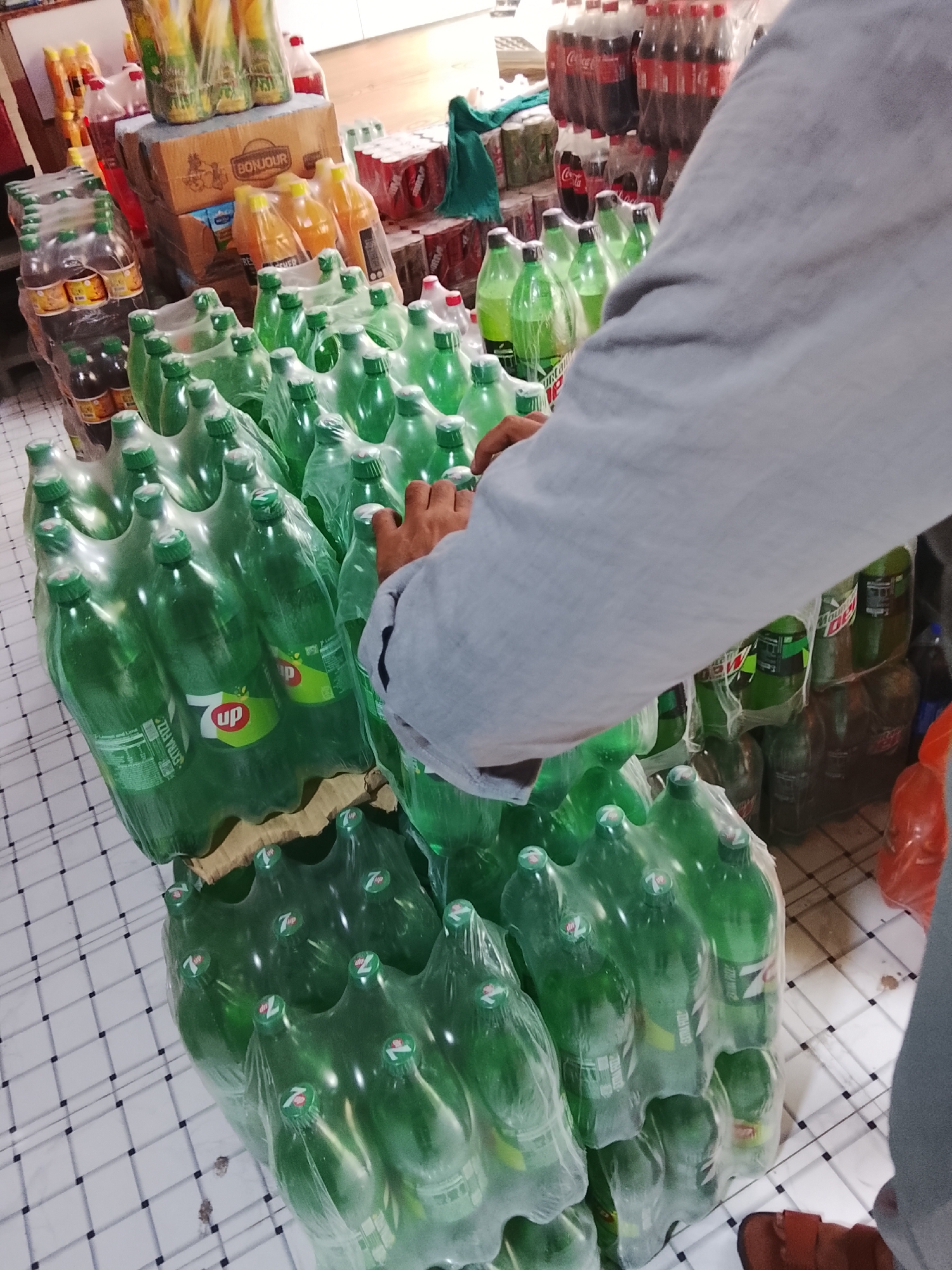 The image shows a cluttered convenience store or wholesale depot, possibly in Gulgasht Colony, Pakistan, judging by the available information.
The scene is dominated by stacks of shrink-wrapped 7-Up and Mountain Dew soda bottles. The bottles are green, and stacked 3-4 layers high. Other beverages, including Coca-Cola, are visible in the background. There are also packages of orange juice. A person wearing a grey shirt is reaching for the green soda bottles.
The floor is tiled with a white tile with black grid lines.