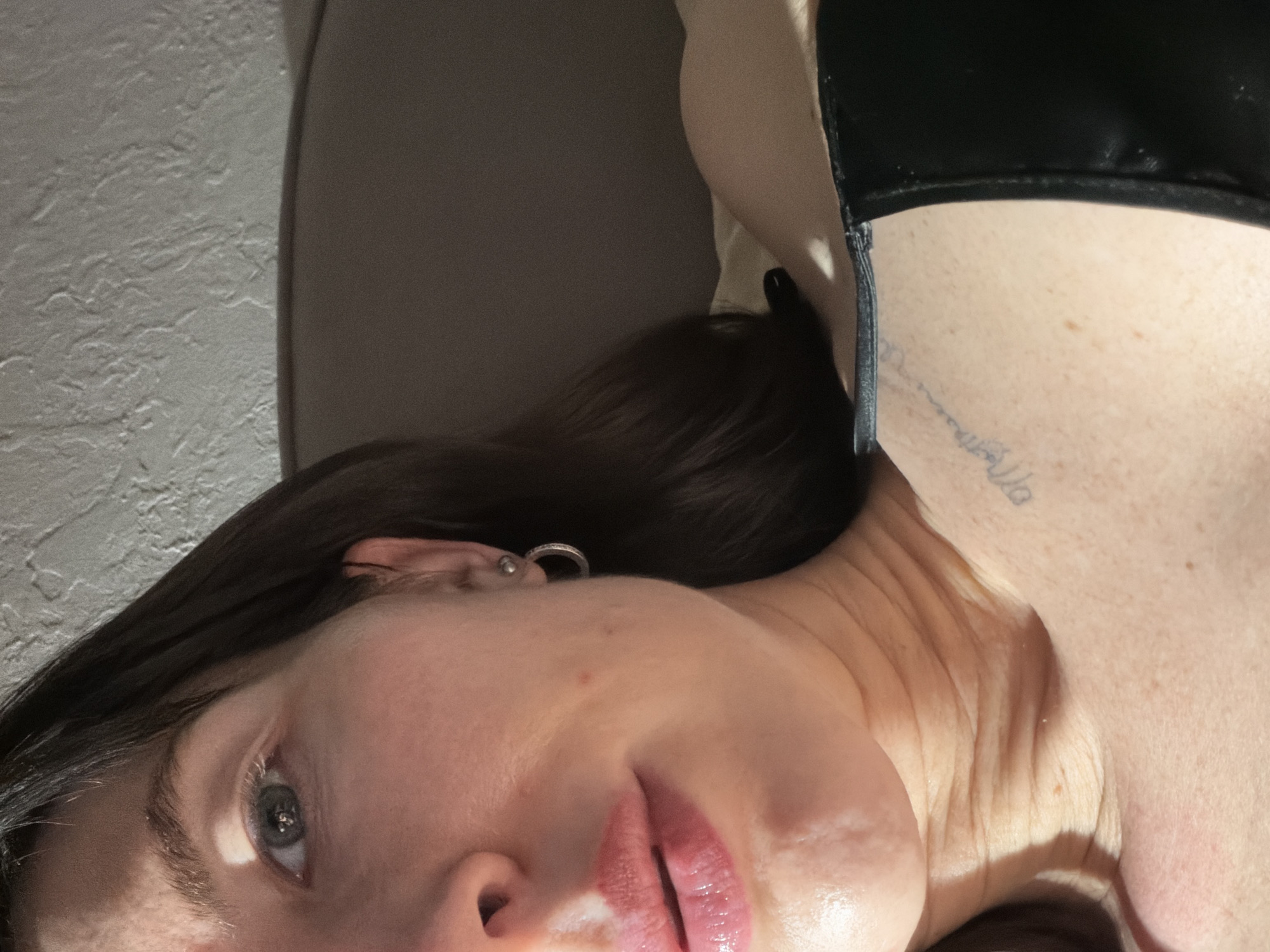 A close-up, high-angle portrait shot features a female subject positioned with her face angled towards the camera. The left side of her face is prominently visible, including her light-colored left eye, the bridge of her nose, and her glossy, full lips. Her left ear is adorned with a small silver hoop earring and a small silver stud. Dark hair is visible along the right side of the frame. Strong, directional lighting, consistent with sunlight, casts significant highlights on the subject's upper left forehead, cheek, and shoulder, creating distinct shadows. The subject's left shoulder and clavicle area are exposed, revealing a faded, script-style tattoo on the upper chest, partially readable as "Magdalena." A dark-colored garment, appearing to be a bra strap or top, is visible along the lower right edge of the frame. The background includes a light-colored, textured wall in the upper right quadrant and a darker, indistinct surface behind the subject's head and right shoulder. The precise location within the city of Perm, Russia, cannot be determined from the visual information provided in this image.