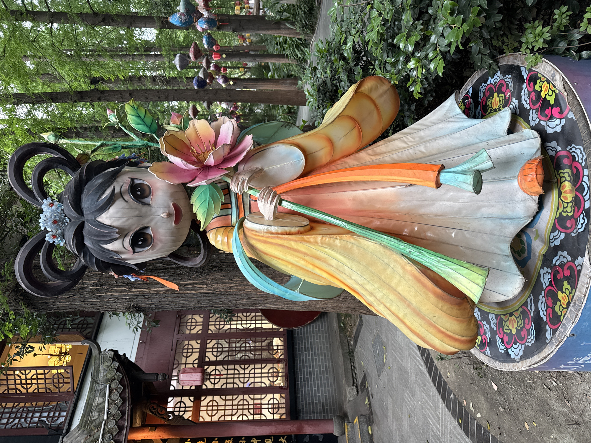 The image shows a large, colorful lantern display of a traditional Chinese character in a park-like setting. The lantern is positioned on a decorated platform with a black and colorful floral pattern. The character is holding a large pink flower and has elaborate black hair ornaments adorned with blue flowers. The lantern's clothing is a mix of yellow, orange, and white hues.
In the background, there are tall trees and foliage, suggesting a natural setting. Further back, a glimpse of traditional Chinese architecture can be seen, including a building with latticed windows and curved rooftops. The scene appears to be in an outdoor location, possibly a garden or park. Based on the request for the location, it is likely situated in the Qingyang District, China.