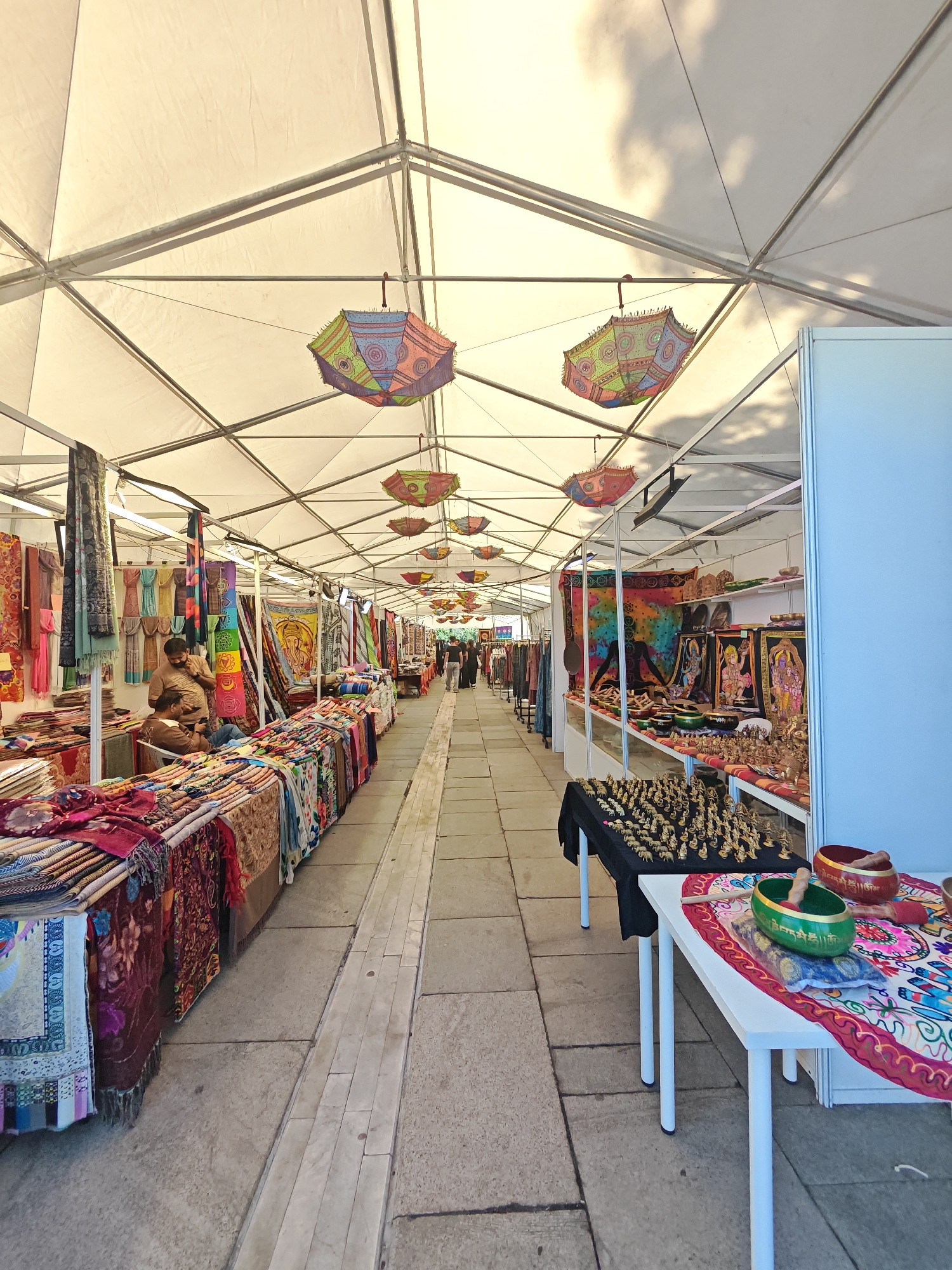 This image depicts an outdoor market stall, likely a temporary setup under a large, white, fabric canopy supported by a metal frame. The market appears to be located in Sofia, Bulgaria, given the presence of traditional Bulgarian textiles and crafts, as well as the distinct architectural style often found in the city's open-air markets.
The stall is filled with various items for sale. On the left side, neatly folded textiles, possibly scarves or cloths with intricate patterns and vibrant colors, are displayed on tables. Some of these textiles feature traditional Bulgarian embroidery or motifs. A man in a brown t-shirt and another man in a light brown shirt are standing and interacting near the textile display.
Further down the aisle, the market continues with more stalls. Hanging from the canopy are colorful, decorative umbrellas, adding a festive atmosphere to the scene. These umbrellas are patterned with geometric designs and bright colors.
On the right side of the image, there are more display tables. One table is covered with a black cloth and showcases numerous small, golden figurines, possibly of deities or decorative statues. Adjacent to this, a white table displays Tibetan singing bowls of various sizes and colors, along with a mallet. The singing bowls are decorated with intricate inscriptions. Behind these tables, a stall displays framed artwork, likely religious or traditional in theme, and other decorative items.
The ground is paved with large, light-colored stone tiles. In the background, the market extends further, with more stalls and people browsing. The overall impression is of a bustling, colorful, and culturally rich market environment. The lighting suggests it is daytime, with natural light filtering through the canopy, and shadows from clouds are visible on the fabric.