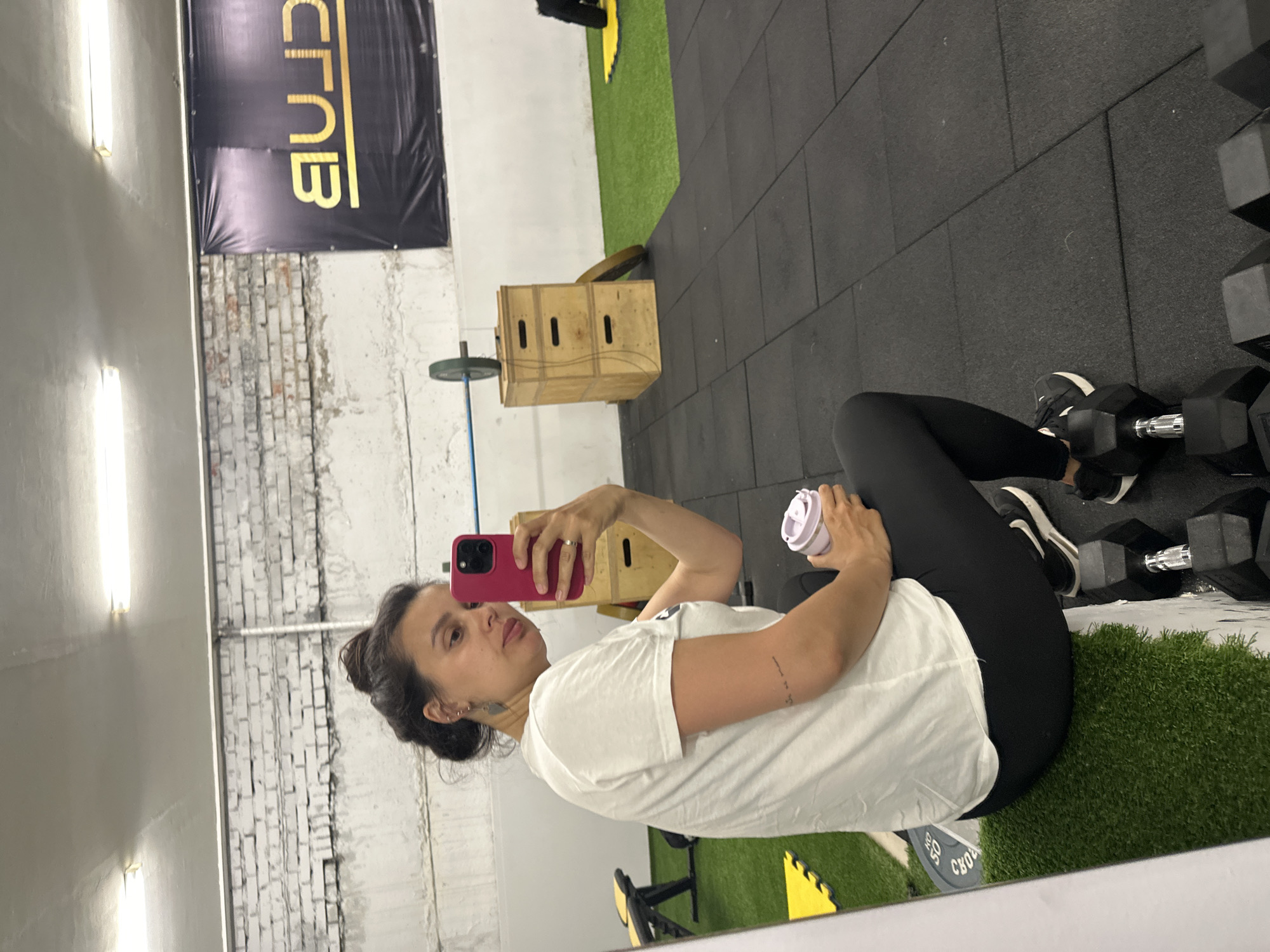A woman is taking a selfie in a gym. She is seated on an area of artificial turf, next to a rack of dumbbells. She is wearing black leggings, black and white sneakers, and a white t-shirt. She is holding a pink cell phone in her right hand and a white shaker bottle in her left. The gym has a variety of workout equipment including weight plates and a plyometric box. A banner with the word "CLUB" written vertically hangs on the wall. The gym floor is covered with black rubber mats and patches of artificial turf. The location is assumed to be in Chaikovsky, Russia.