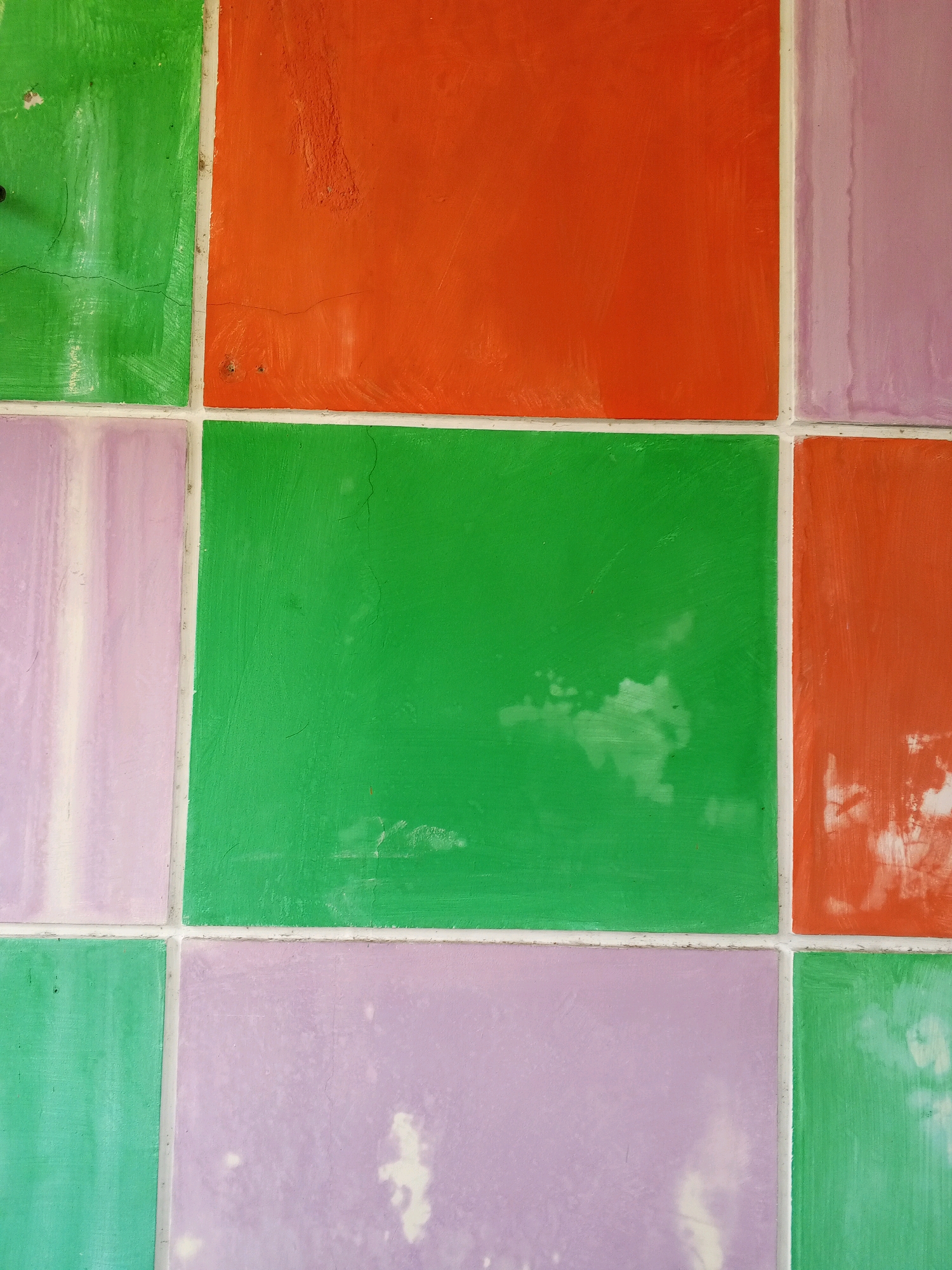 The image is a close-up, top-down view of a tiled wall in Condongcatur, Indonesia. The wall is comprised of rectangular tiles in alternating colors of green, orange, and purple, laid out in a grid pattern. The tiles appear to be made of concrete or a similar material and show signs of wear, including minor cracks and some patchy discoloration, particularly on the purple and orange tiles. The grout lines between the tiles are visible and are a light, neutral color, likely white or off-white. The lighting is even, suggesting an overcast day or an indoor setting with diffused light. No people or specific objects are depicted, focusing solely on the texture and color of the tiled surface.