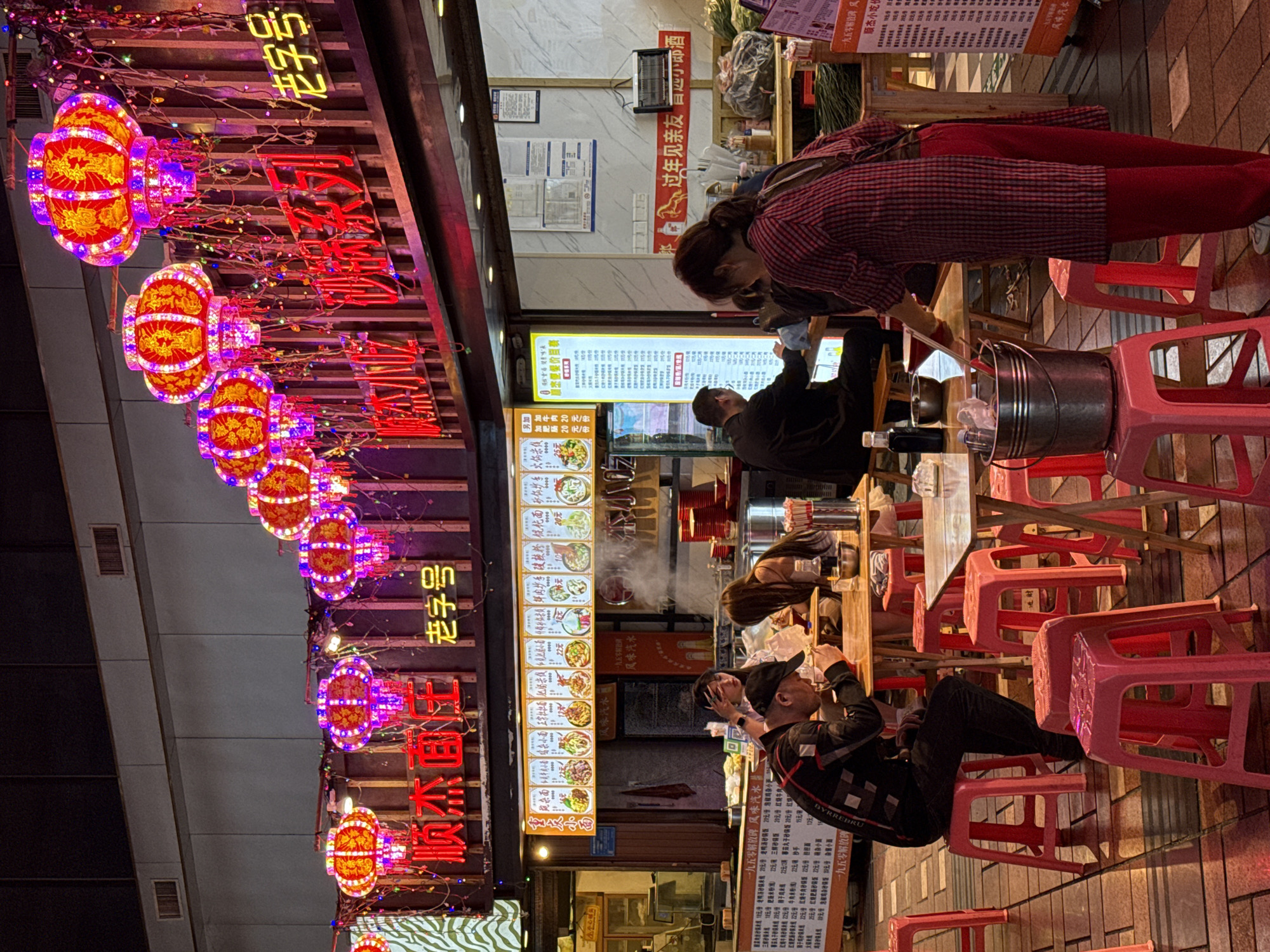Here's a detailed description of the image:
**Scene:** The image depicts an outdoor restaurant in what appears to be the Yuzhong District of China, judging by the Chinese characters and general ambiance. It is nighttime, and the restaurant is brightly lit with neon signs and decorative lanterns.
**People:** Several people are visible in the image:
* A woman in a red and black plaid shirt is standing near a table and appears to be using chopsticks to get food.
* A man in a suit is seated at the table and appears to be looking at a phone.
* A woman with long hair is seated at the table and appears to be eating food.
* A man is sitting on a small stool and drinking something.
**Objects:** The following objects are visible:
* A row of brightly colored lanterns and neon signs adorns the restaurant's storefront.
* A food menu is displayed on a board with Chinese characters and food images.
* A table and small pink stools are arranged for diners.
* Utensils, dishes, and what appears to be a metal bucket with utensils are on the table.
**Actions and Interactions:**
* People are eating, drinking, and using phones.
* There seems to be a group of people dining together.
**Environment:**
* The setting is a bustling outdoor area with food stalls and restaurants.
* The ground appears to be a brick or stone-paved surface.
* The scene is well-lit with a mix of neon signs and street lighting.